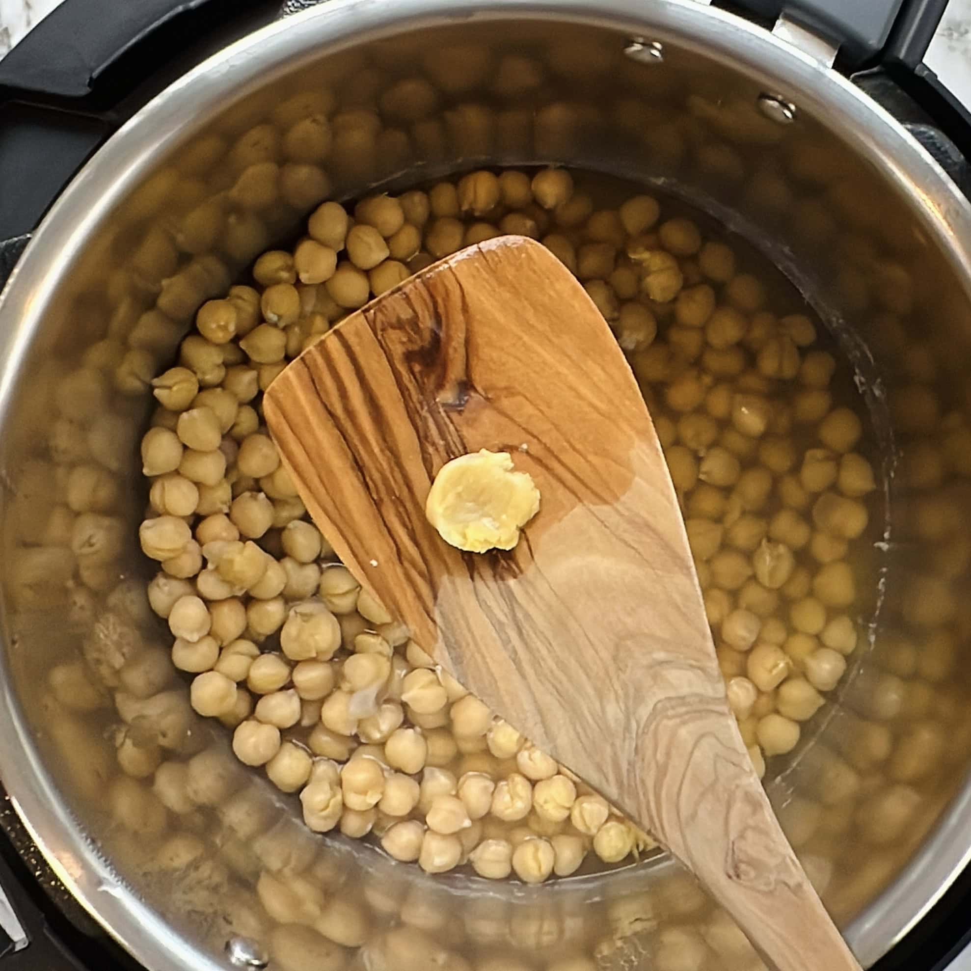 A wooden spatula holds a single peeled chickpea over a pot filled with unpeeled chickpeas submerged in water.