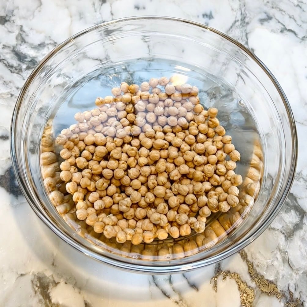 A glass bowl filled with dried chickpeas soaking in water sits on a marble countertop.