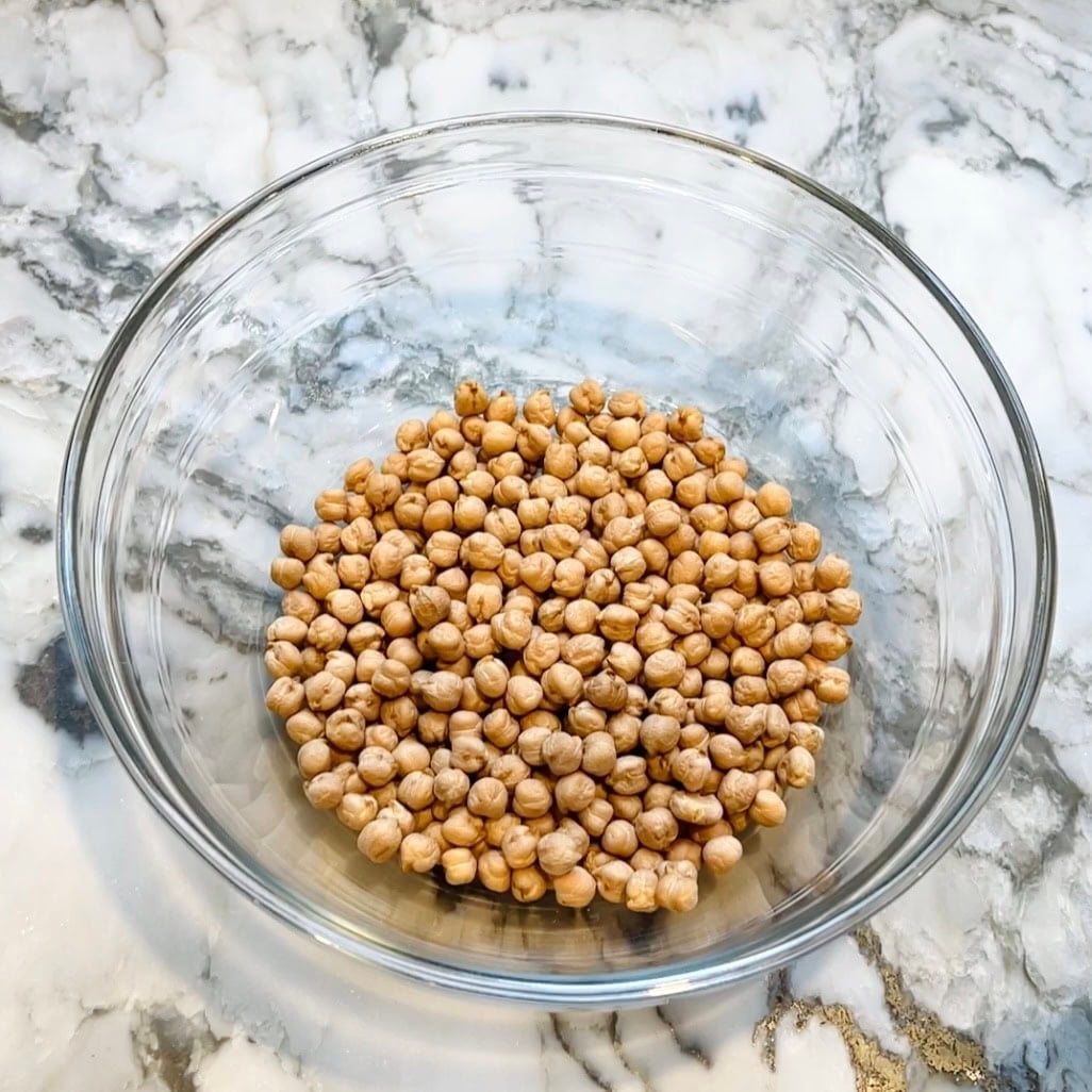 A clear glass bowl containing dried chickpeas sits on a marble countertop.
