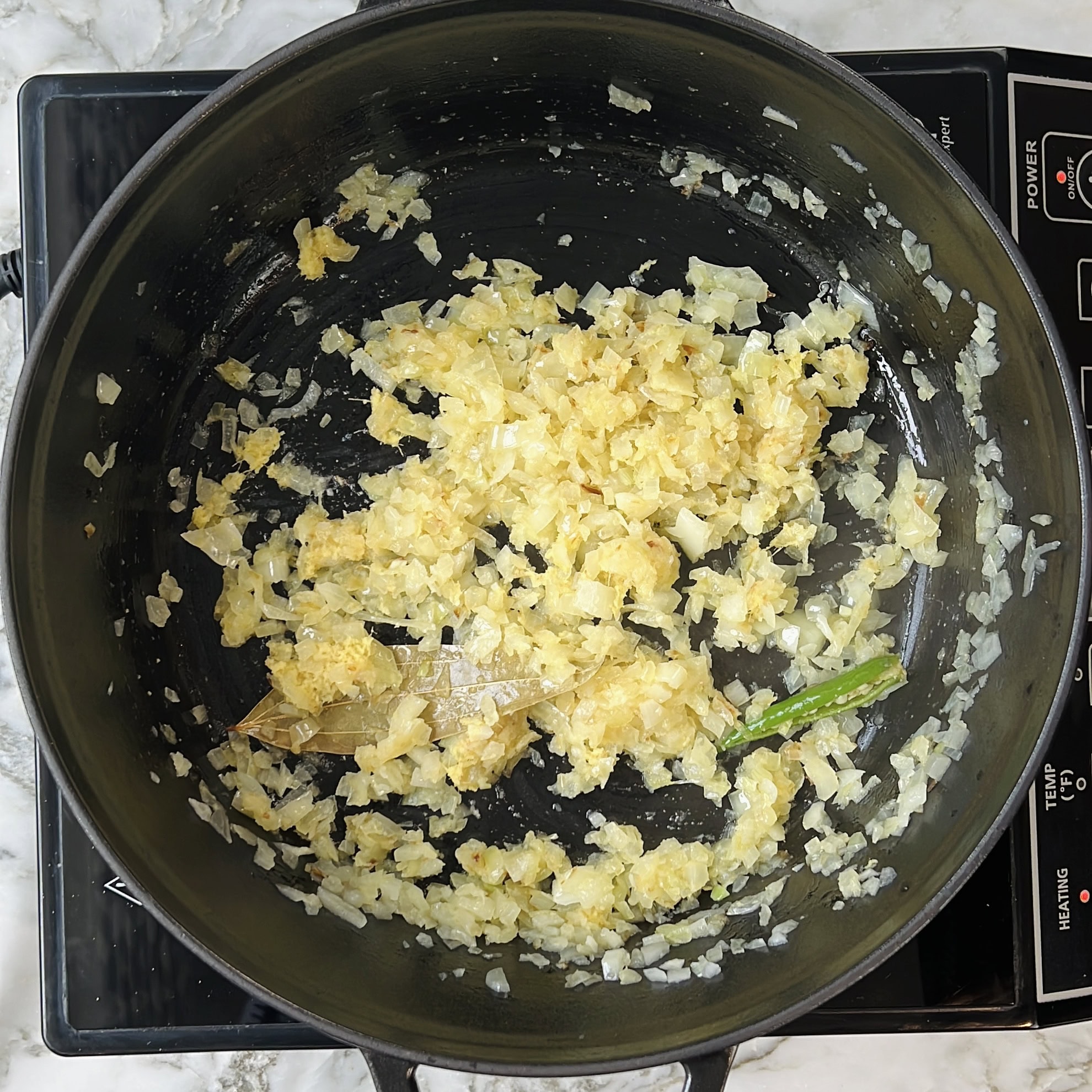 Chopped onions, garlic, ginger, a bay leaf, and a green chili are sautéing in a large black pot on an electric stovetop.