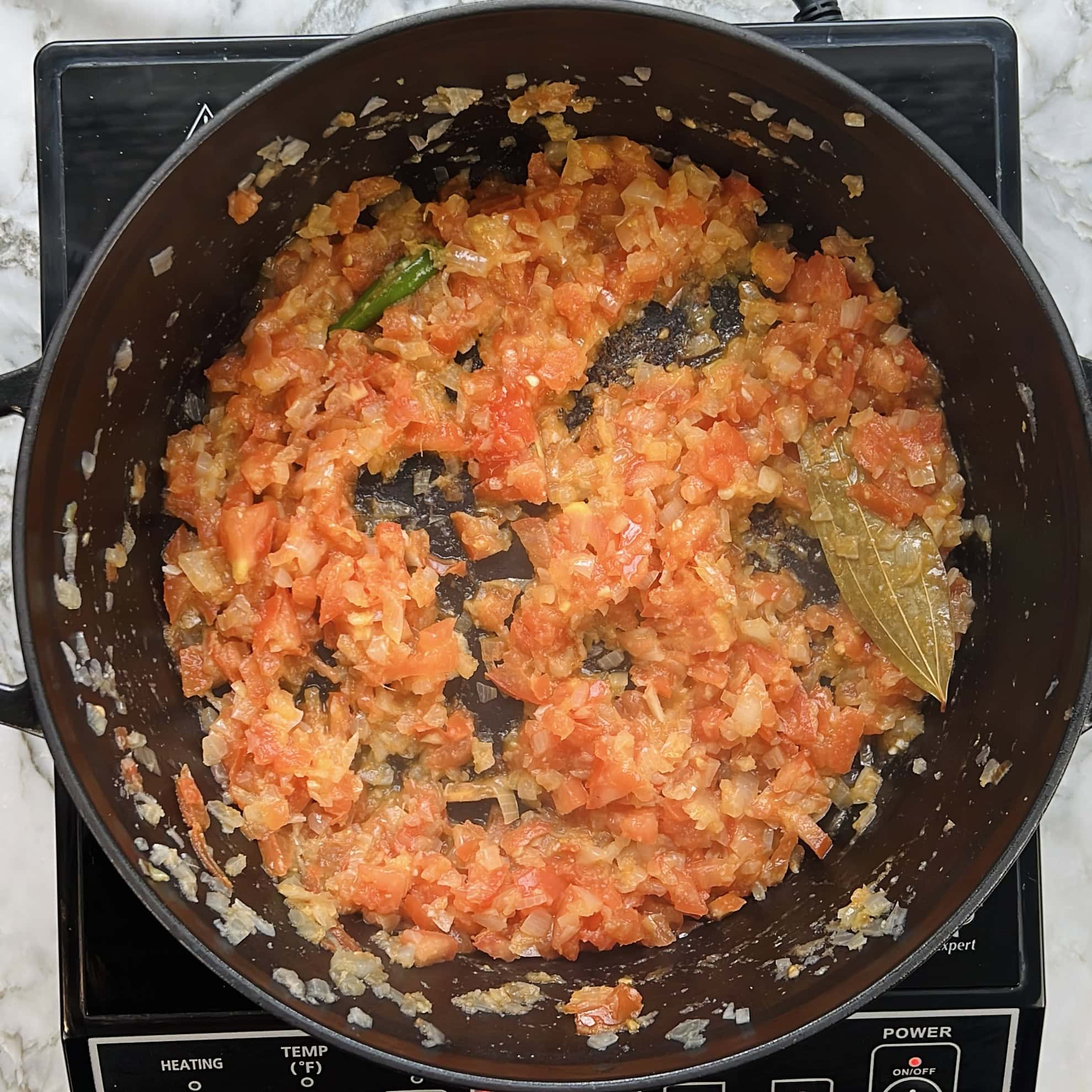 Chopped onions, tomatoes, green chili, and a bay leaf are being sautéed in a black pan on an electric stovetop.
