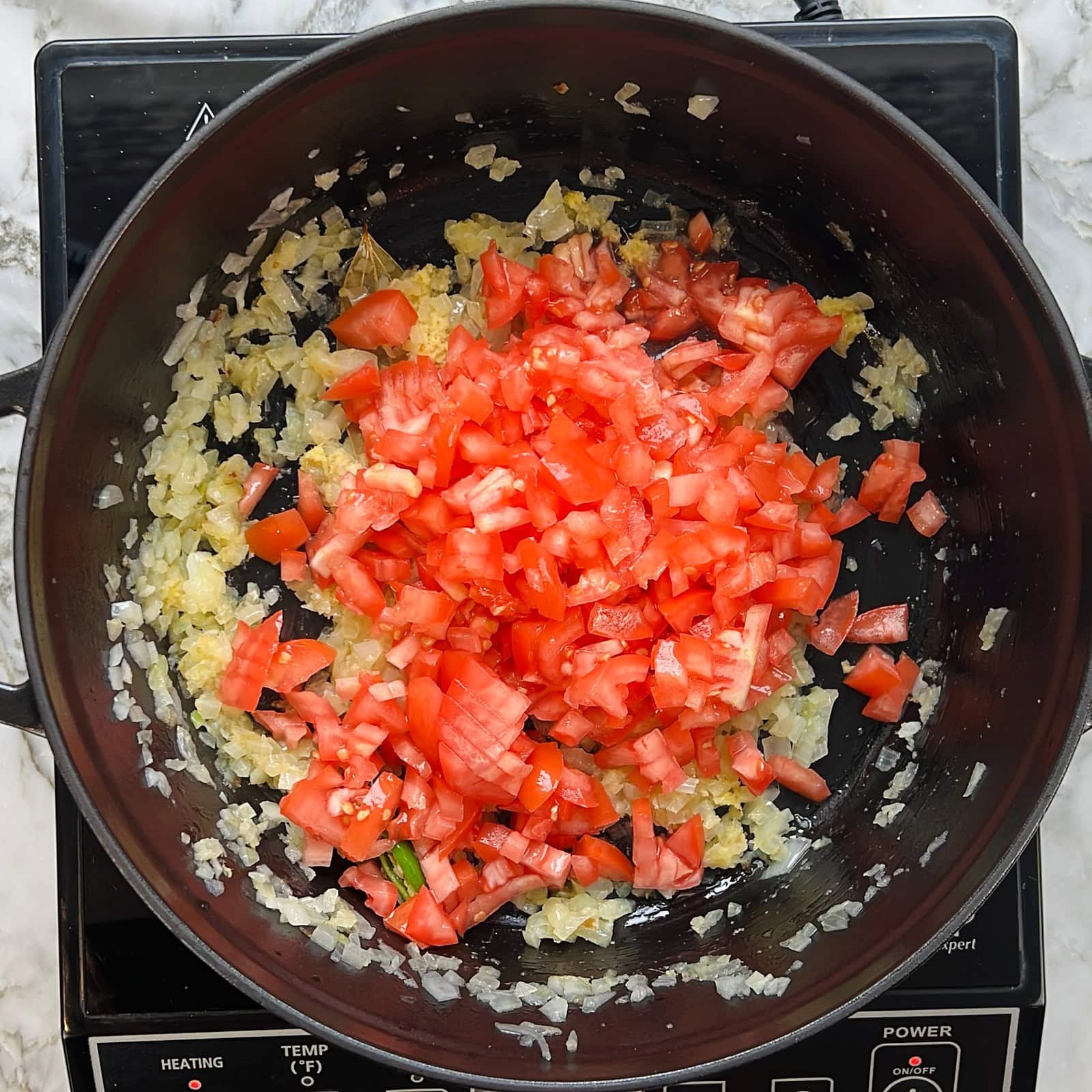 Chopped onions and tomatoes cooking in a black pan on an electric stove.