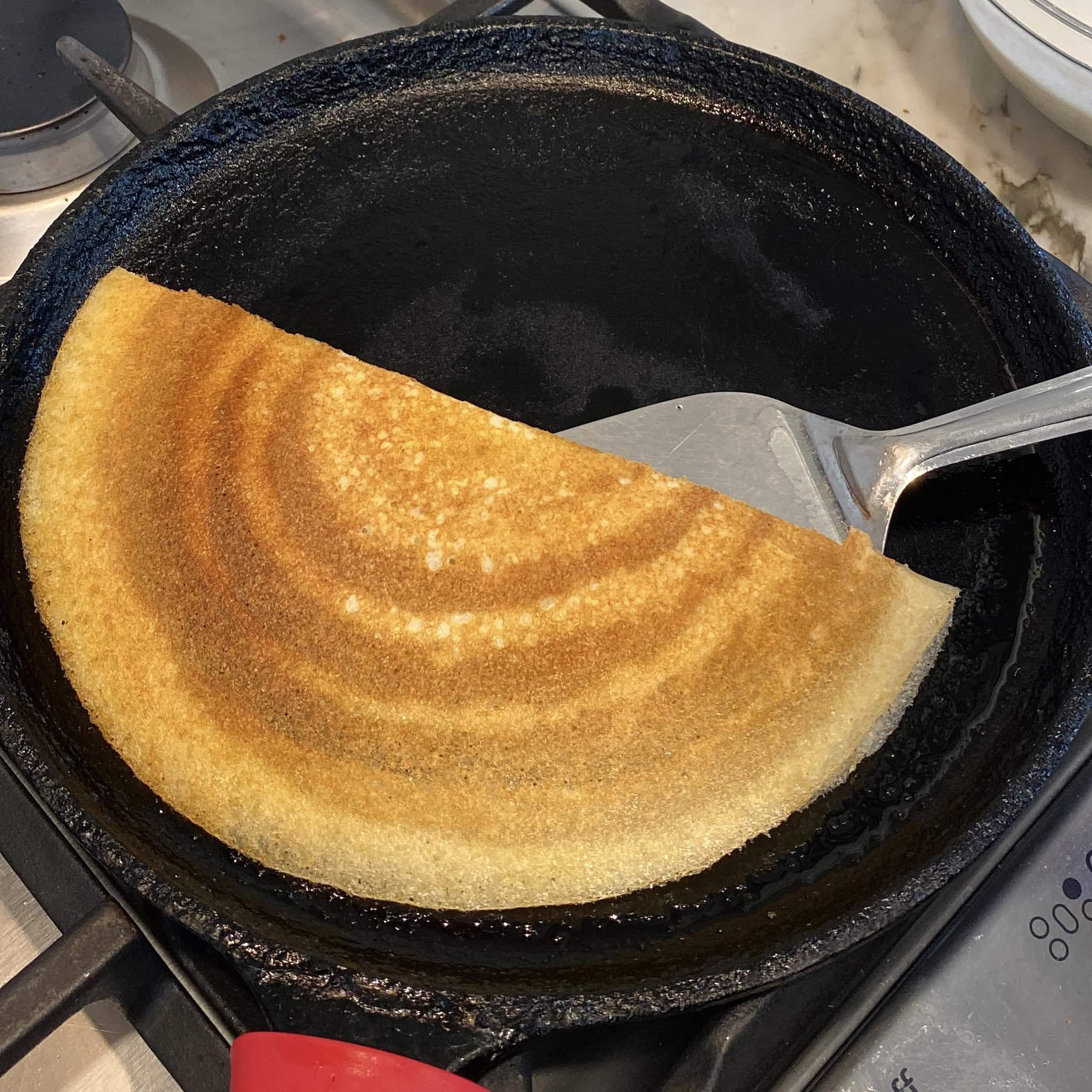 A golden-brown dosa being lifted with a spatula from a black cast iron pan on a stovetop.