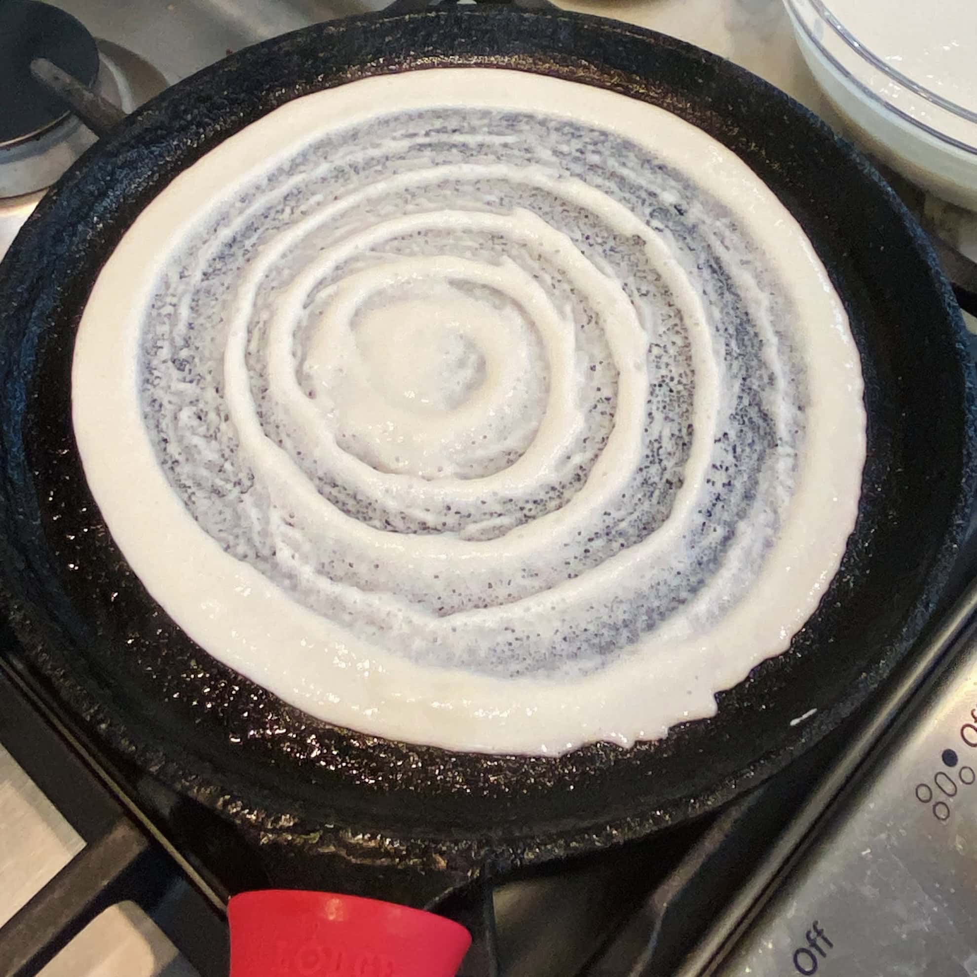 A dosa being cooked on a cast iron pan over a stove, with circular batter patterns visible on the surface.