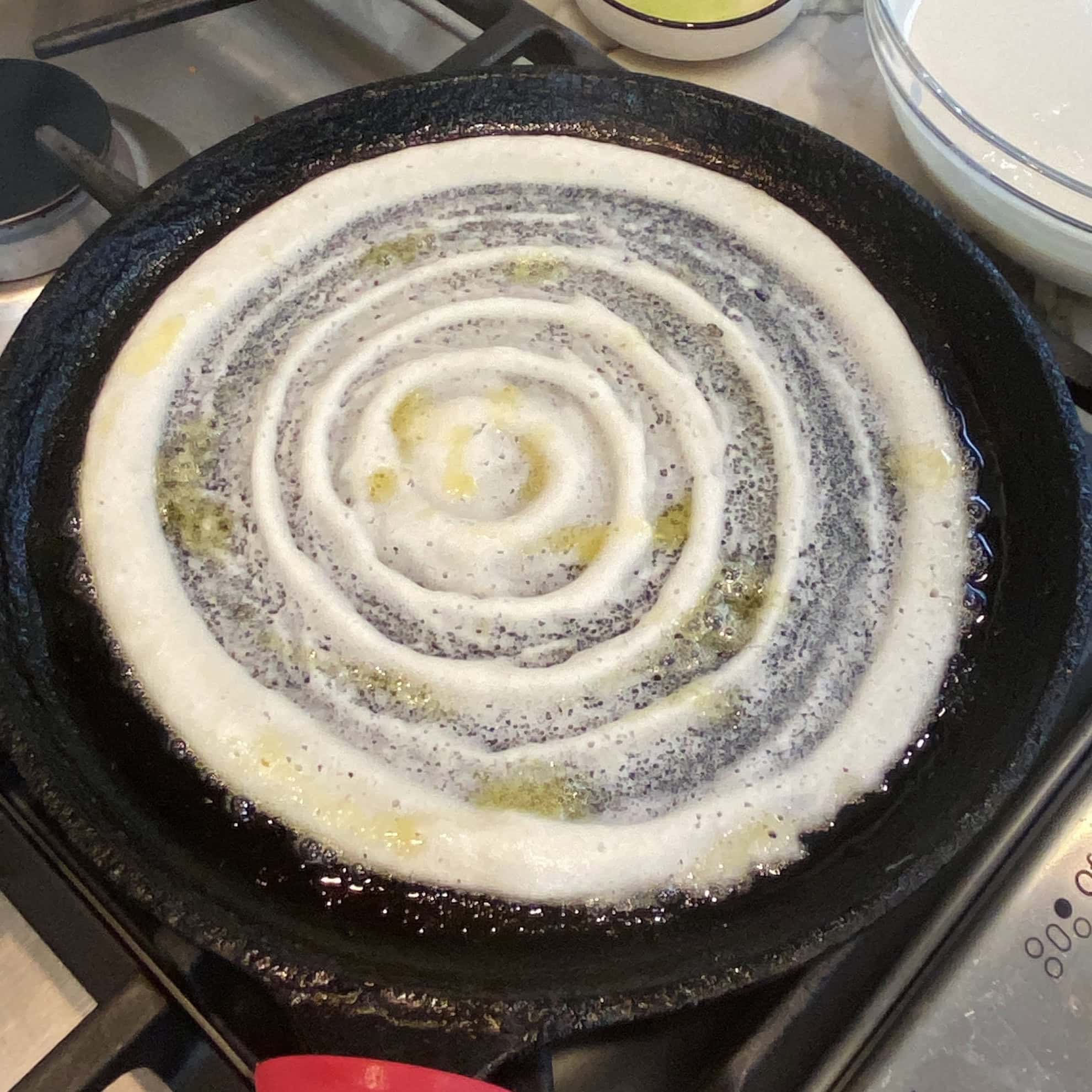 A dosa is being cooked on a black cast iron pan over a stove, with visible spiral patterns and some oil spread on the surface.
