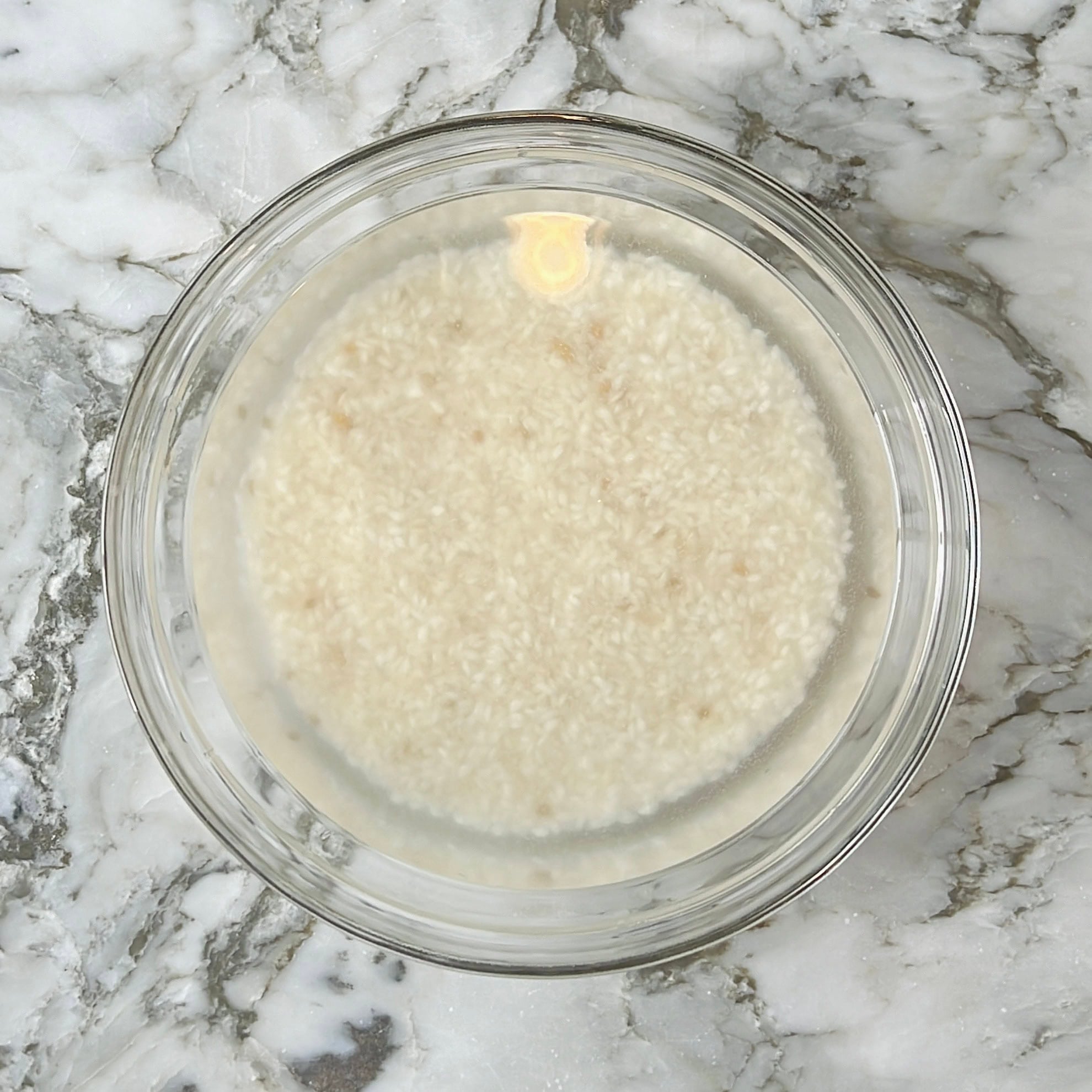 A glass bowl filled with rice soaking in water, placed on a marble countertop.