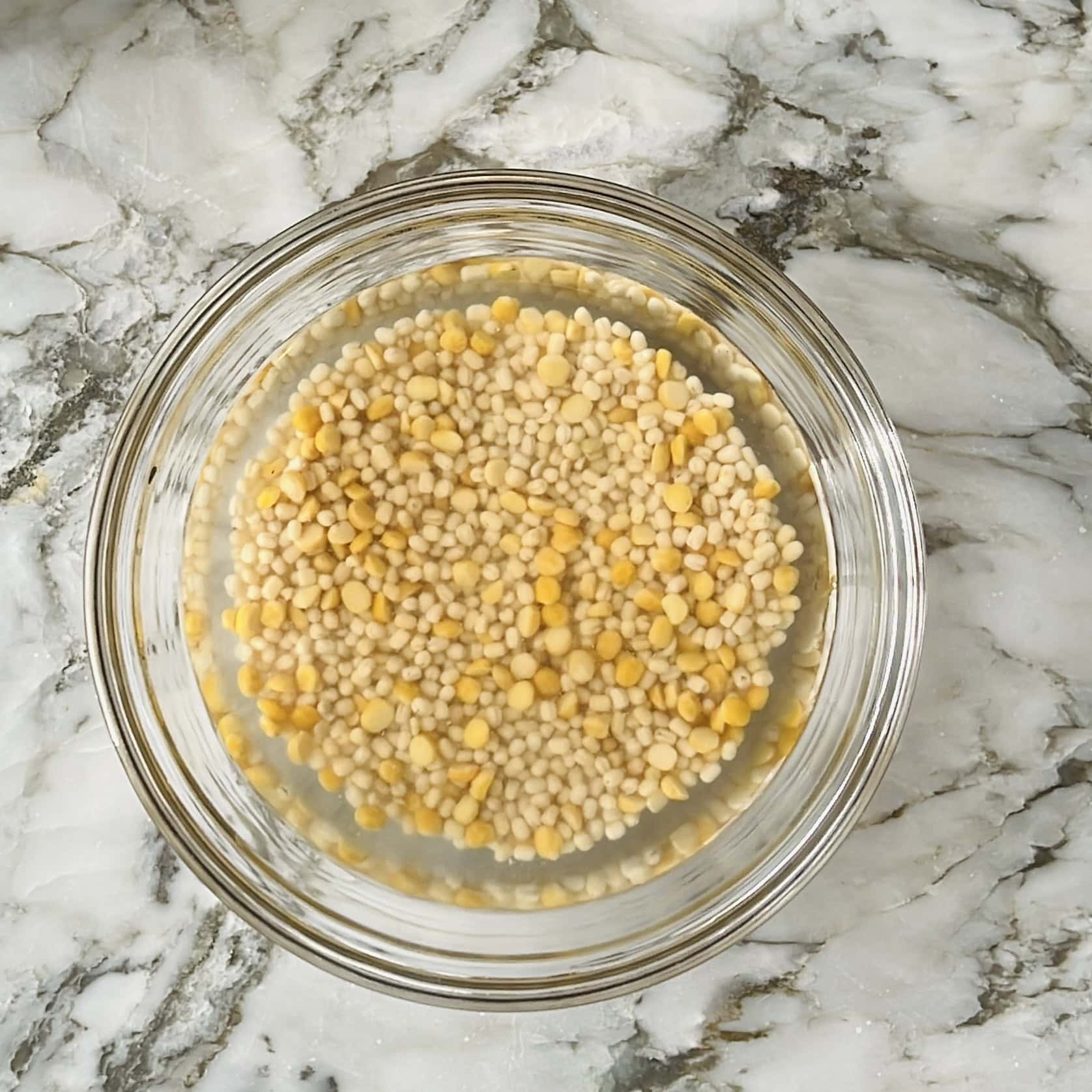 A glass bowl filled with yellow and white lentils soaking in water, placed on a marble countertop.