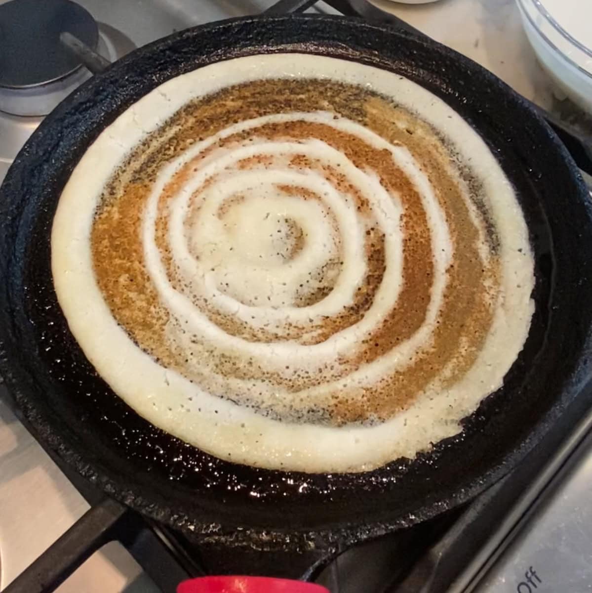 A dosa is cooking on a hot black skillet, showing a spiral pattern of batter on a stovetop.