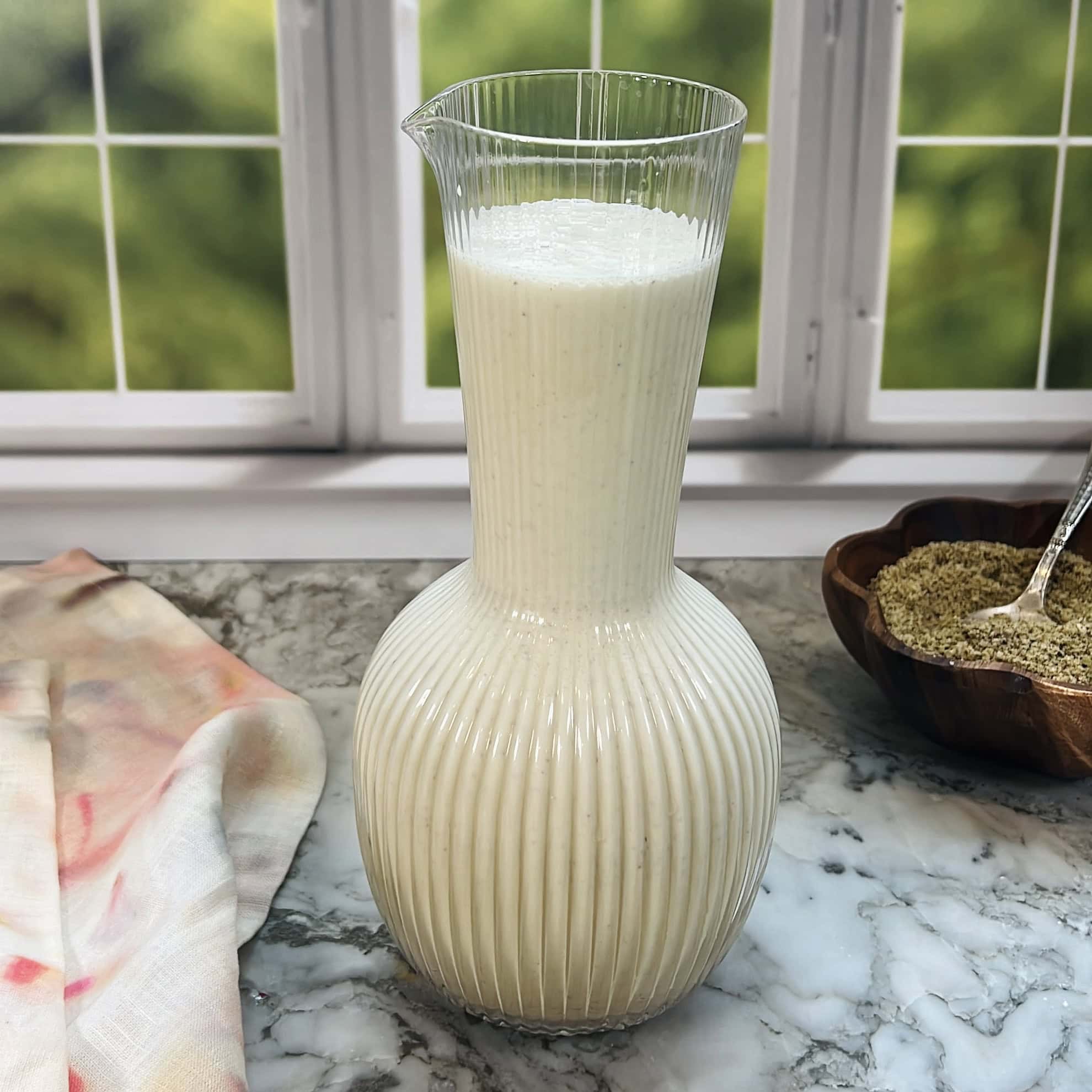 A ribbed glass pitcher filled with a creamy white liquid sits on a marble countertop near a window, with a folded cloth and a wooden bowl of powder beside it.