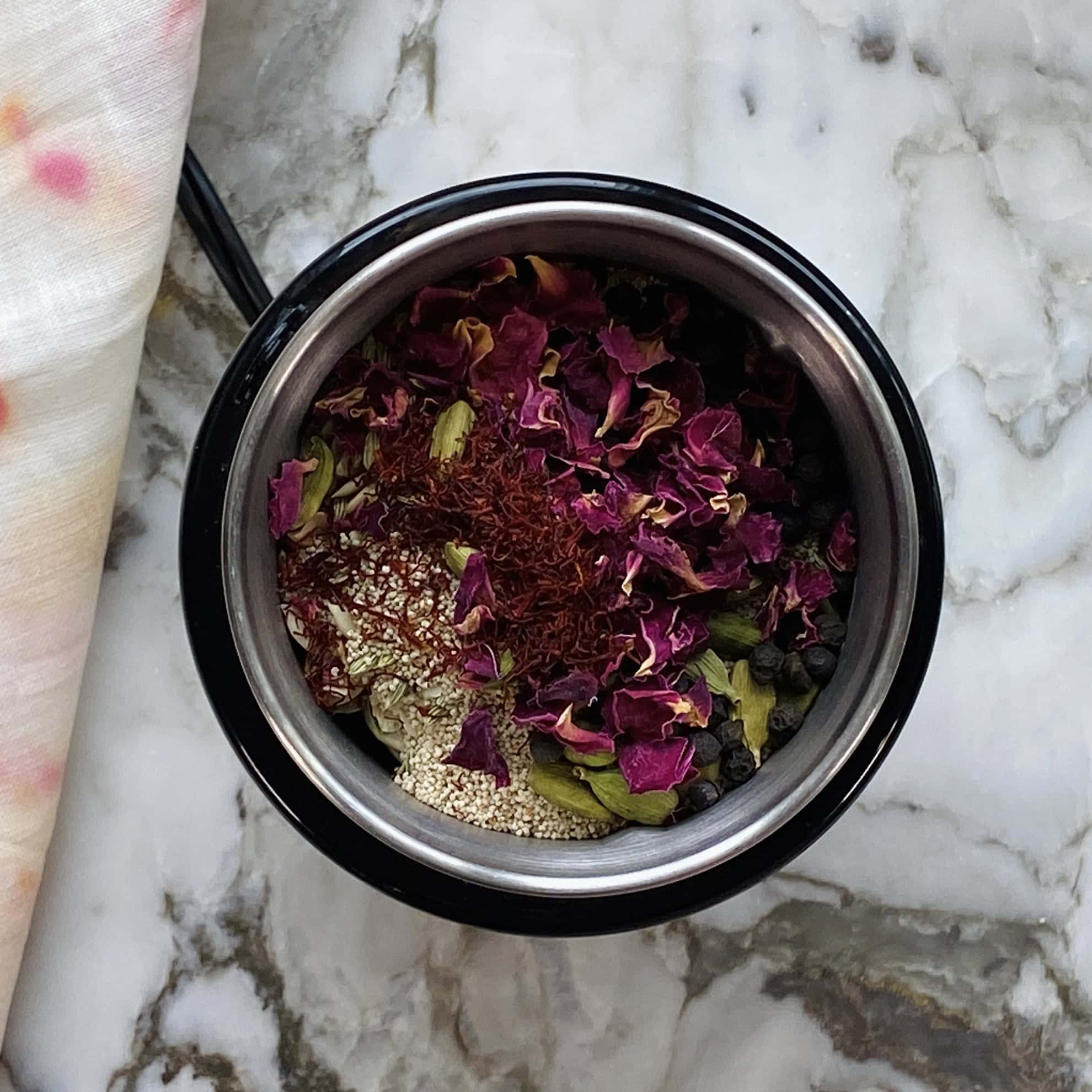 A metal bowl filled with rose petals, saffron threads, cardamom pods, and other spices sits on a marble surface.