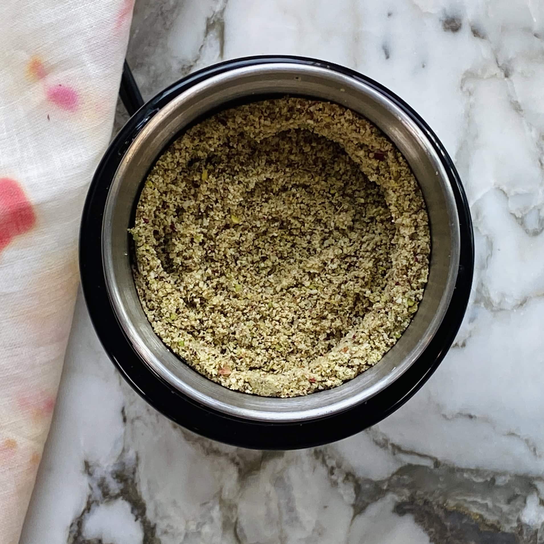A stainless steel grinder bowl containing ground nut mixture sits on a marble countertop next to a white cloth with pink and orange stains.
