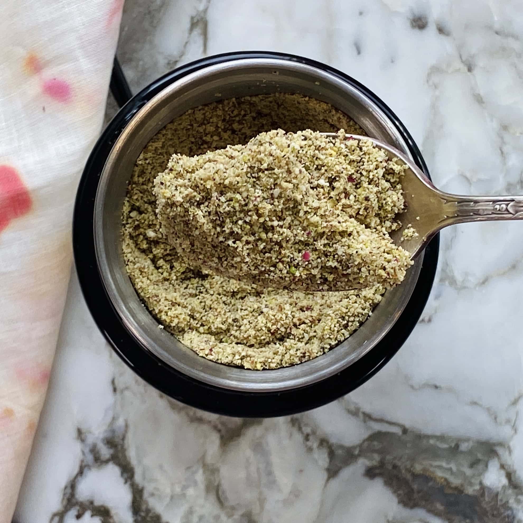 A metal spoon scoops a coarse, greenish-brown powder from a small black bowl on a marble countertop.