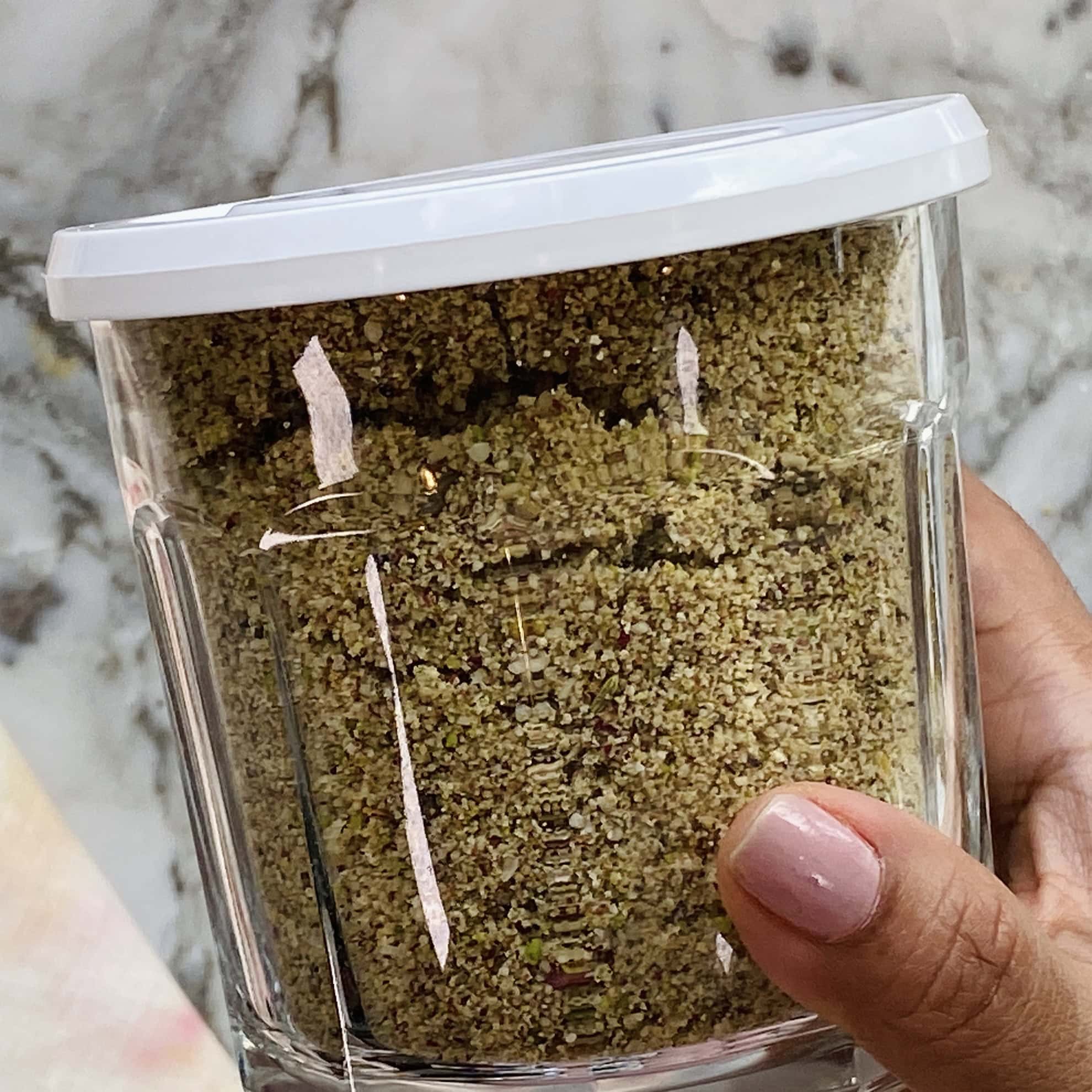 A hand holding a clear plastic container with a white lid, filled with a ground, greenish-brown powder, against a marble background.
