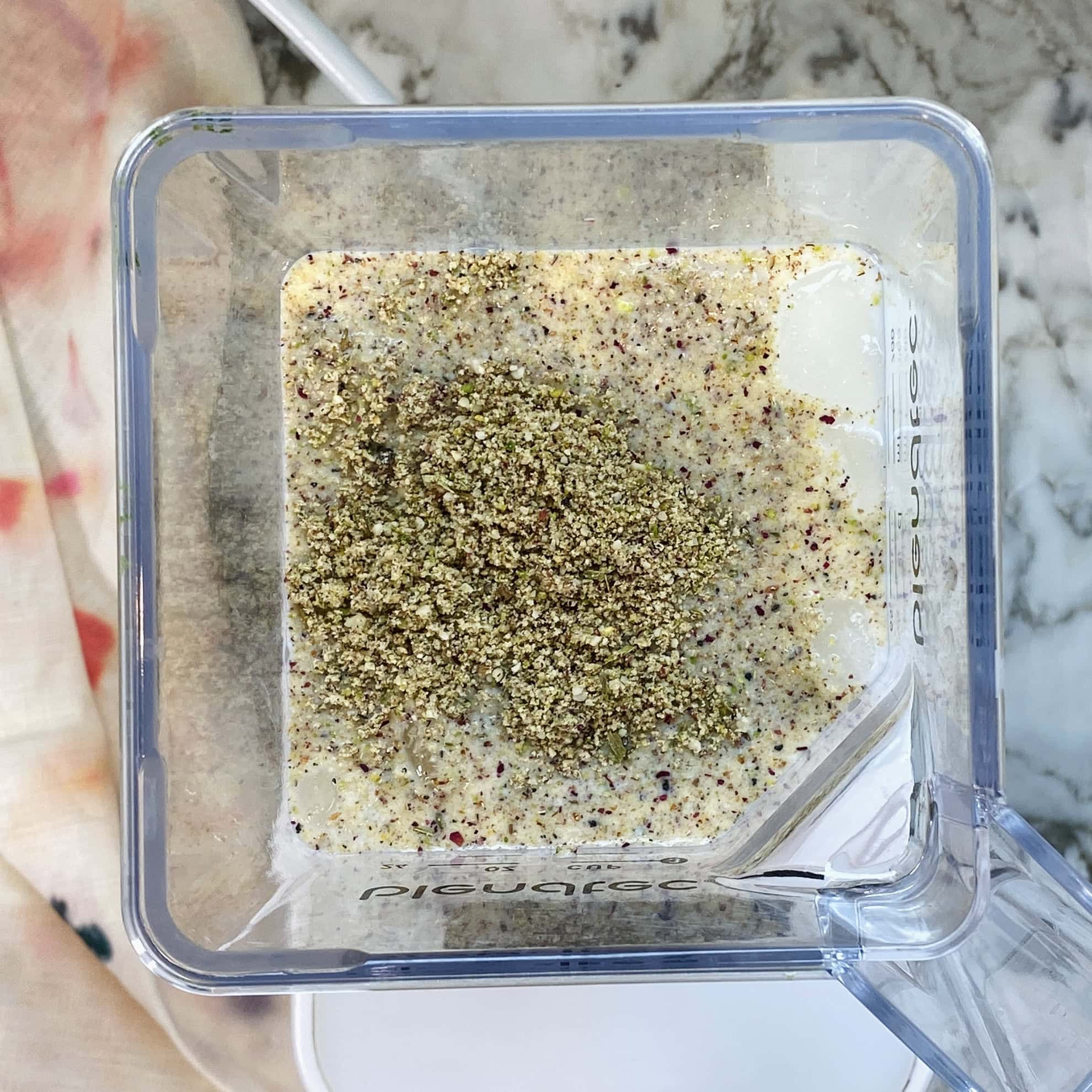 A blender container with milk and a heap of finely ground pistachios inside, sitting on a marble countertop.