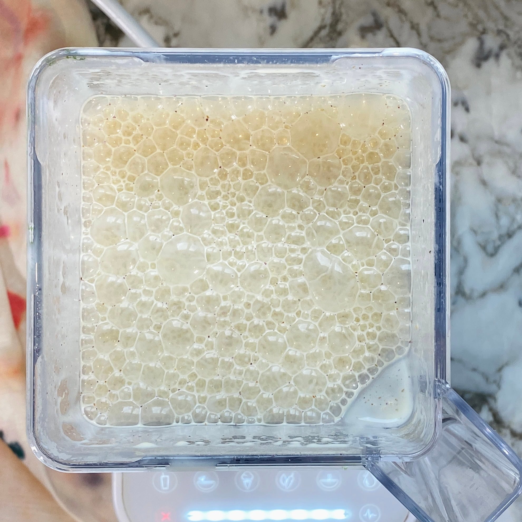 Top-down view of a blender pitcher containing a frothy, light beige liquid with bubbles on the surface, placed on a kitchen countertop.