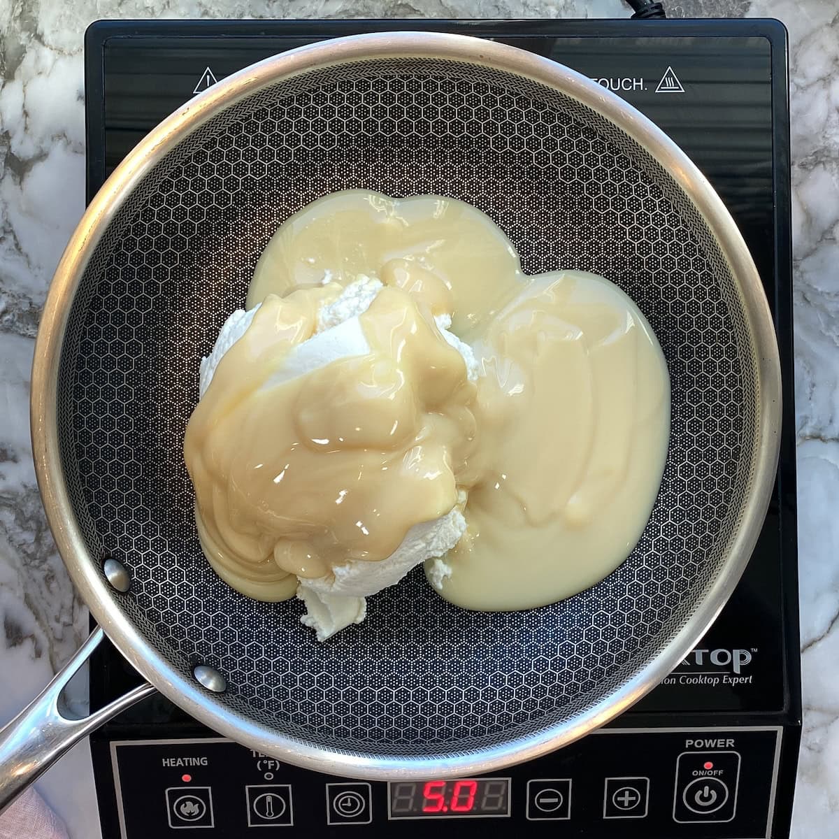 A frying pan on an induction cooktop contains cream cheese and sweetened condensed milk, ready to be heated and mixed.