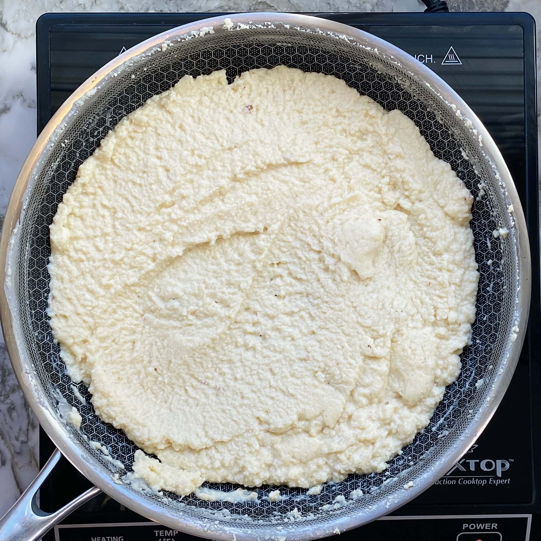 A metal skillet with a layer of white, grainy mixture spread evenly over its surface sits on an induction cooktop.