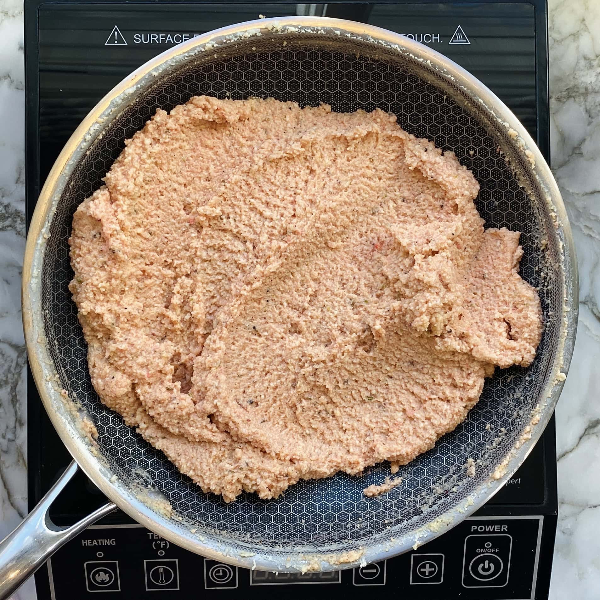 A stainless steel pan on an induction cooktop contains a spread of finely ground pinkish mixture, possibly being cooked or prepared on a marble countertop.