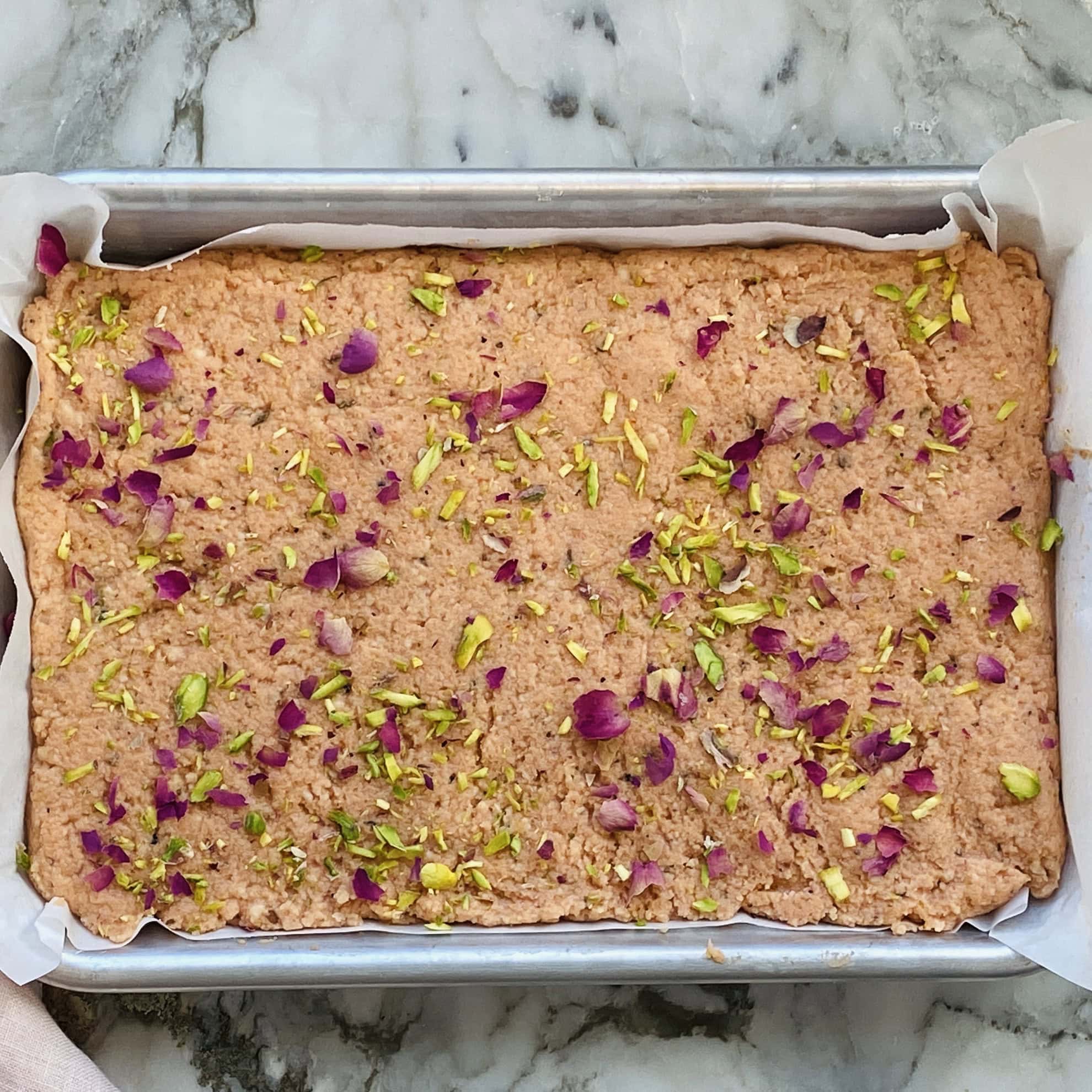 A rectangular baking pan lined with parchment paper, filled with a textured pink dessert topped with chopped pistachios and dried rose petals on a marble surface.