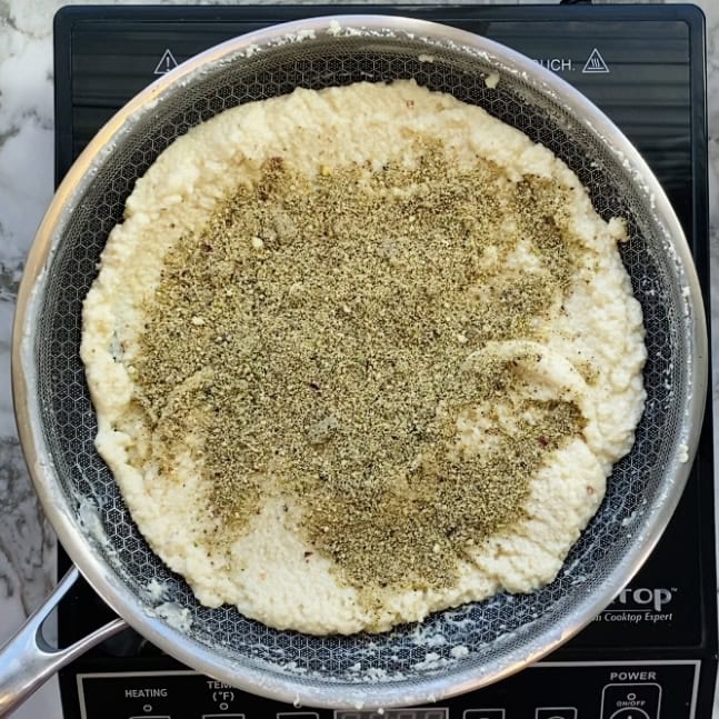 A pan on a stovetop contains a cooking mixture topped with a layer of ground spices or herbs.
