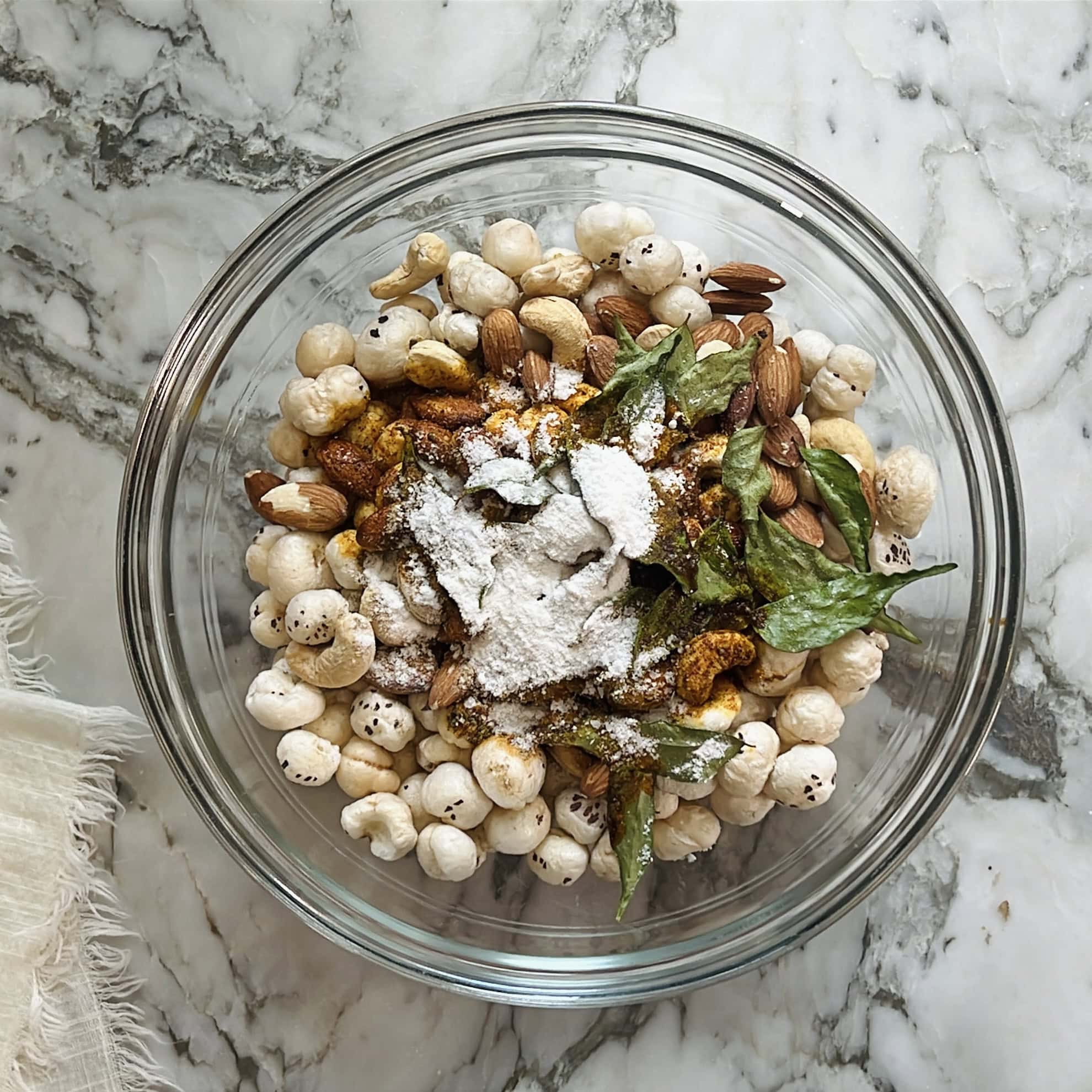 sugar, salt and chat masala added to roasted makahan and nuts in a bowl.