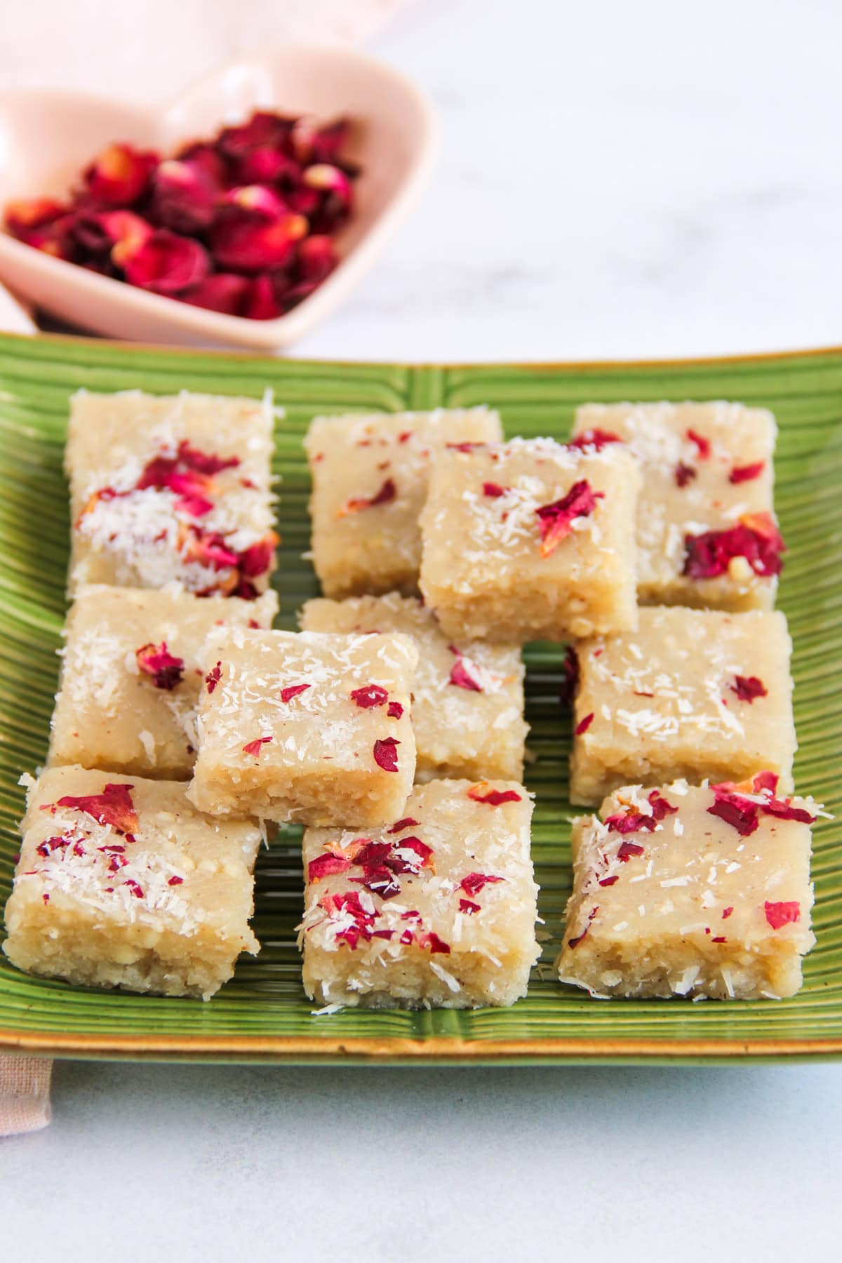 kaju makhana burfi garnished with coconut and dried rose petals on a green platter. 