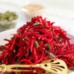 A plate of shredded apple, beet, and carrot salad garnished with seeds and herbs, with a gold serving fork beside it.