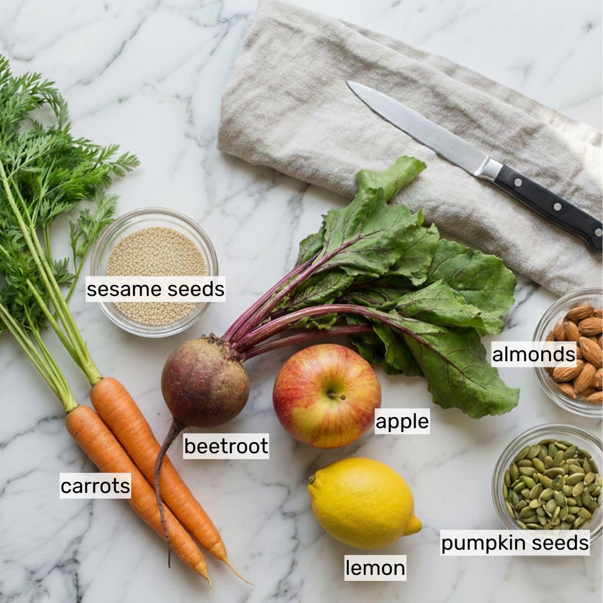 A marble surface with labeled ingredients: carrots, beetroot, apple, lemon, sesame seeds, almonds, pumpkin seeds, a knife, and a cloth napkin.
