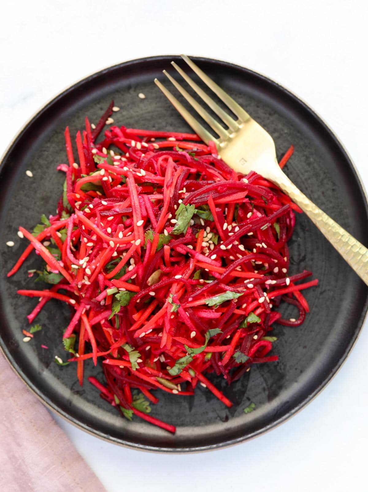 A black plate with a colorful beet and carrot slaw topped with sesame seeds and fresh herbs, next to a gold fork.