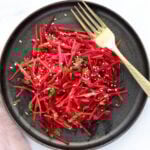A black plate with a salad of thinly sliced red beets and carrots, garnished with sesame seeds and fresh herbs, next to a gold fork.