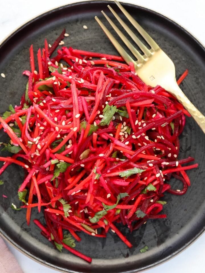 A black plate with a salad of thinly sliced red beets and carrots, garnished with sesame seeds and fresh herbs, next to a gold fork.