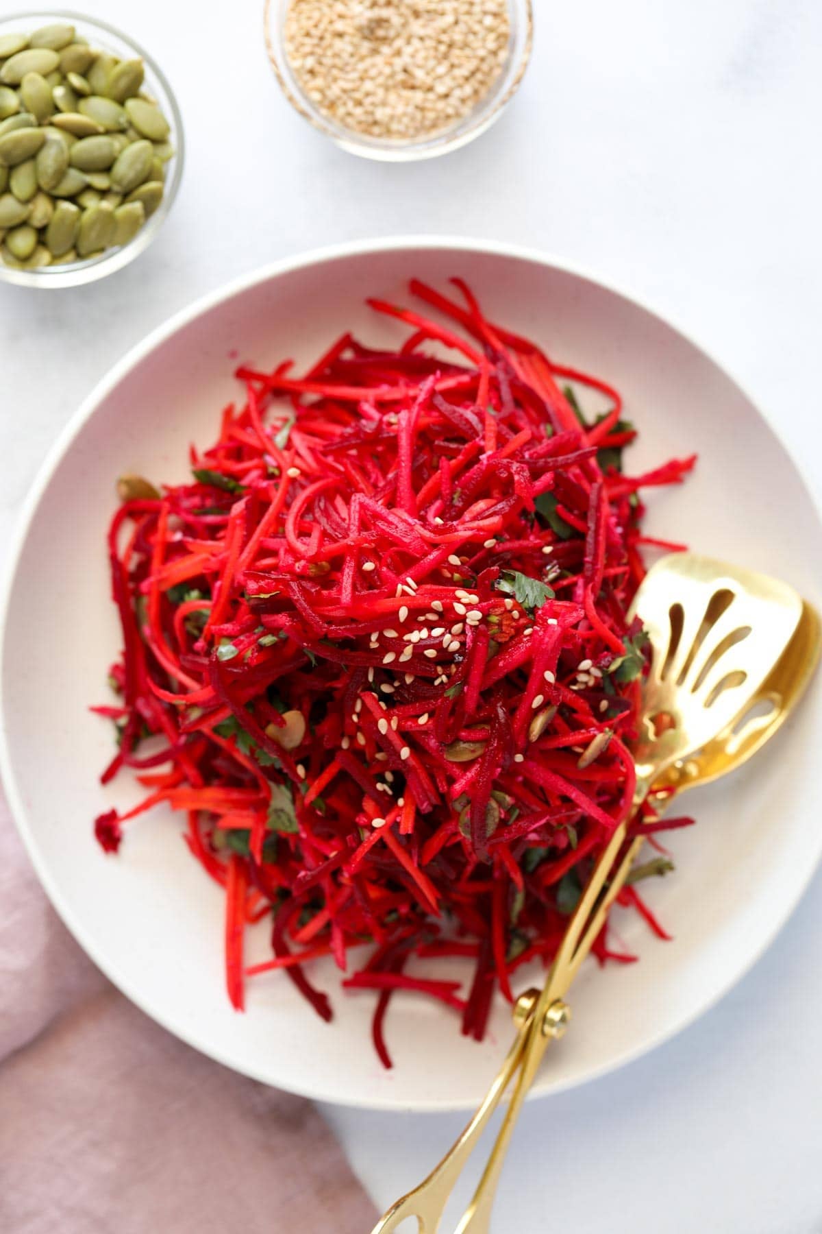A white plate with shredded beet and carrot salad, garnished with sesame seeds and herbs, next to small bowls of pumpkin seeds and sesame seeds. A gold serving utensil is on the plate.