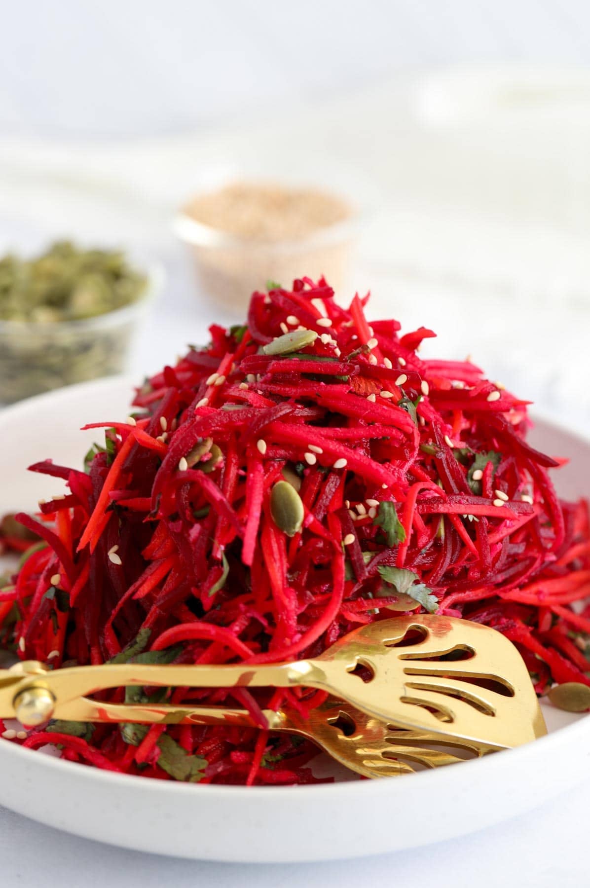 A white plate filled with a shredded beet and carrot salad, garnished with seeds and herbs, with a gold serving fork beside it.