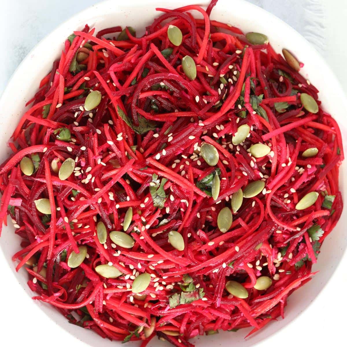 A bowl of shredded beet and carrot salad topped with pumpkin seeds, sesame seeds, and chopped herbs.