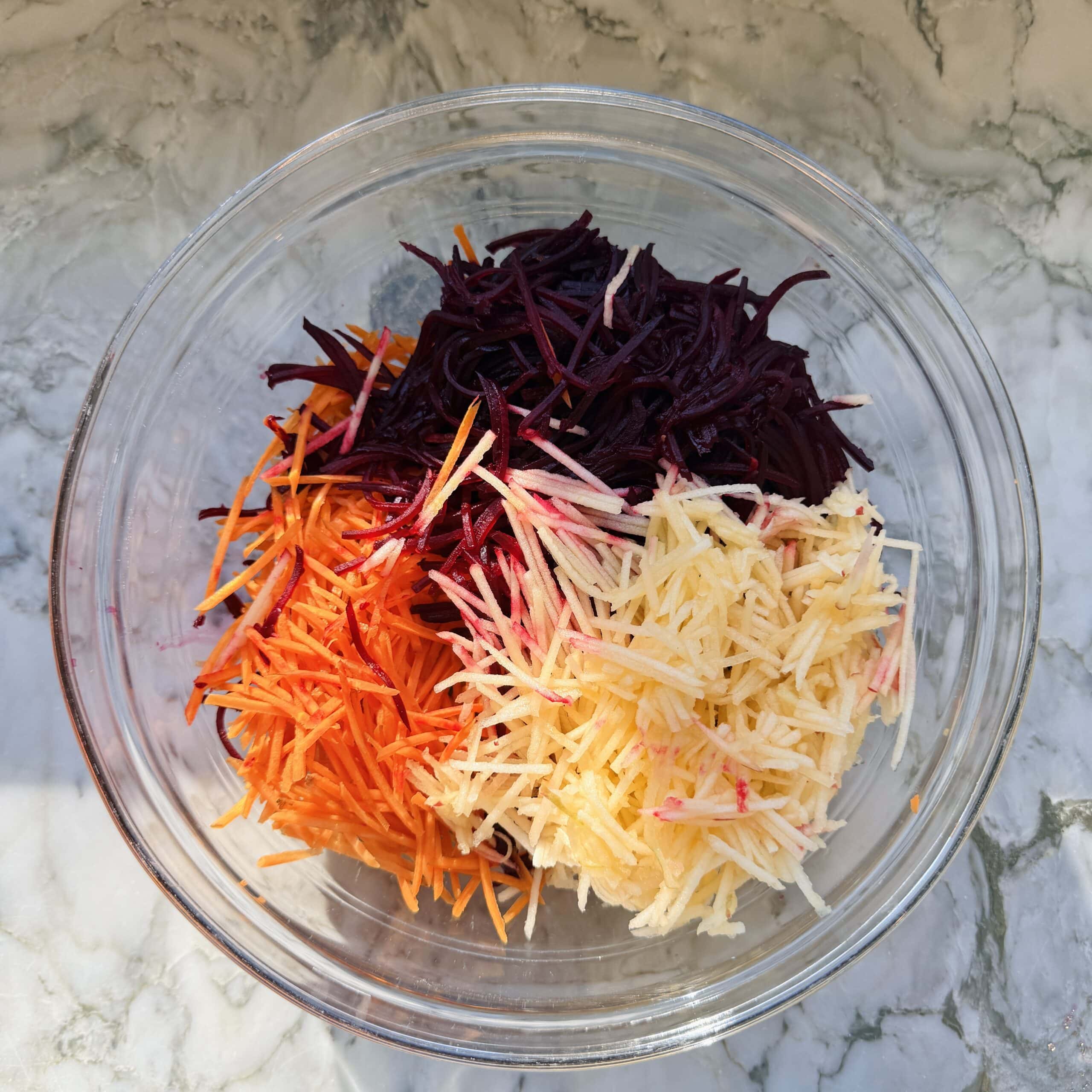 A glass bowl containing separate piles of shredded beetroot, carrot, and white radish on a marble surface.