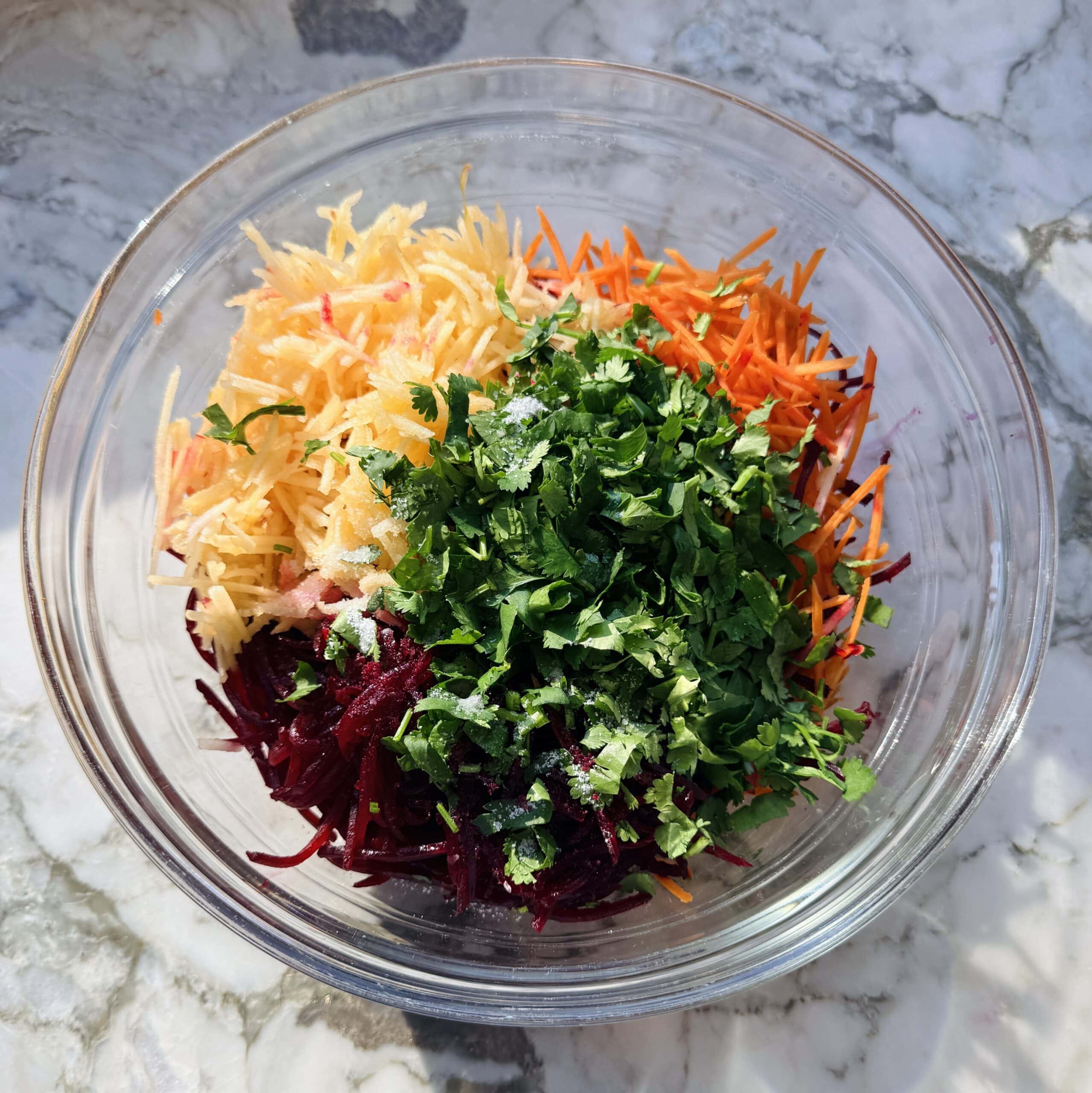 A glass bowl containing shredded beetroot, carrot, and apple topped with chopped fresh cilantro, on a marble surface.