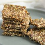 A stack of square-shaped seed crackers with flax, pumpkin, sunflower, and chia seeds on a gray plate.