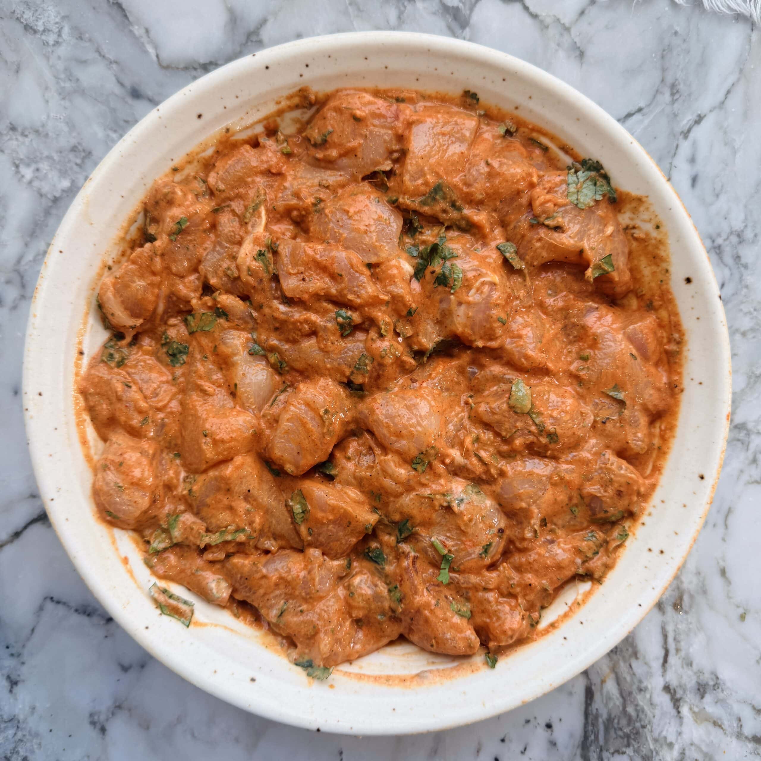 Raw chicken pieces marinated in an orange-red sauce with herbs, arranged in a round white dish on a marble surface.
