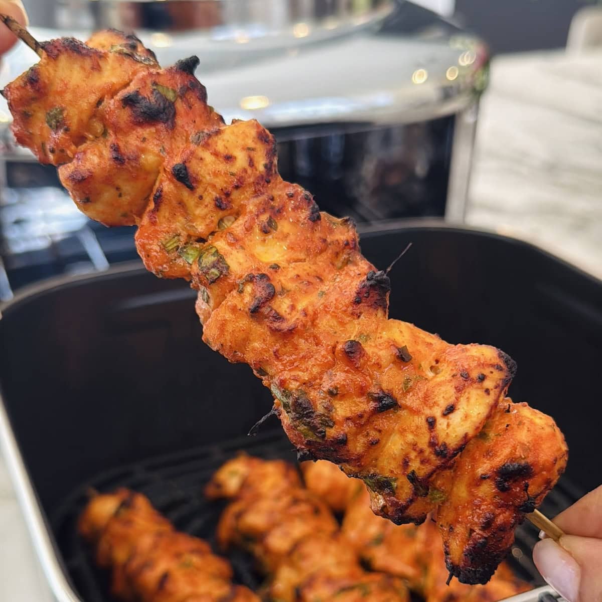 A skewer of grilled, seasoned chicken pieces held above an air fryer, with another skewer visible inside the appliance.