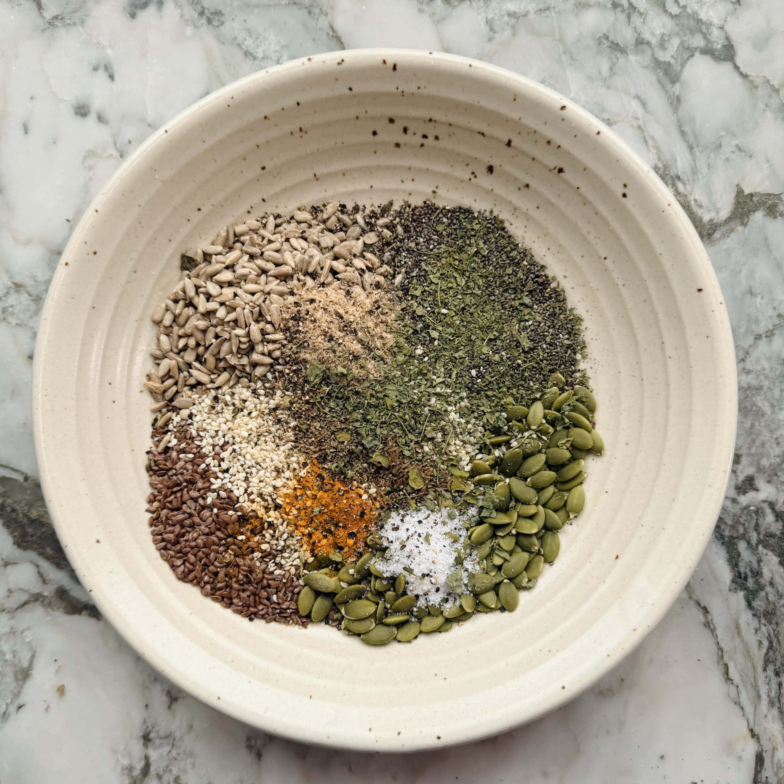 A white bowl containing assorted seeds and spices, including sunflower seeds, pumpkin seeds, flaxseeds, sesame seeds, herbs, salt, pepper, and turmeric, on a marble surface.