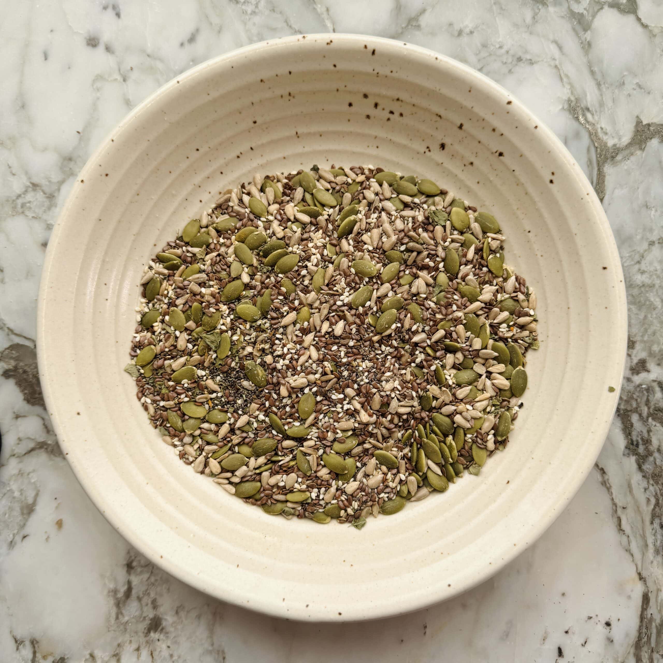 A ceramic bowl containing a mix of seeds, including pumpkin, sunflower, and flax seeds, placed on a marble surface.