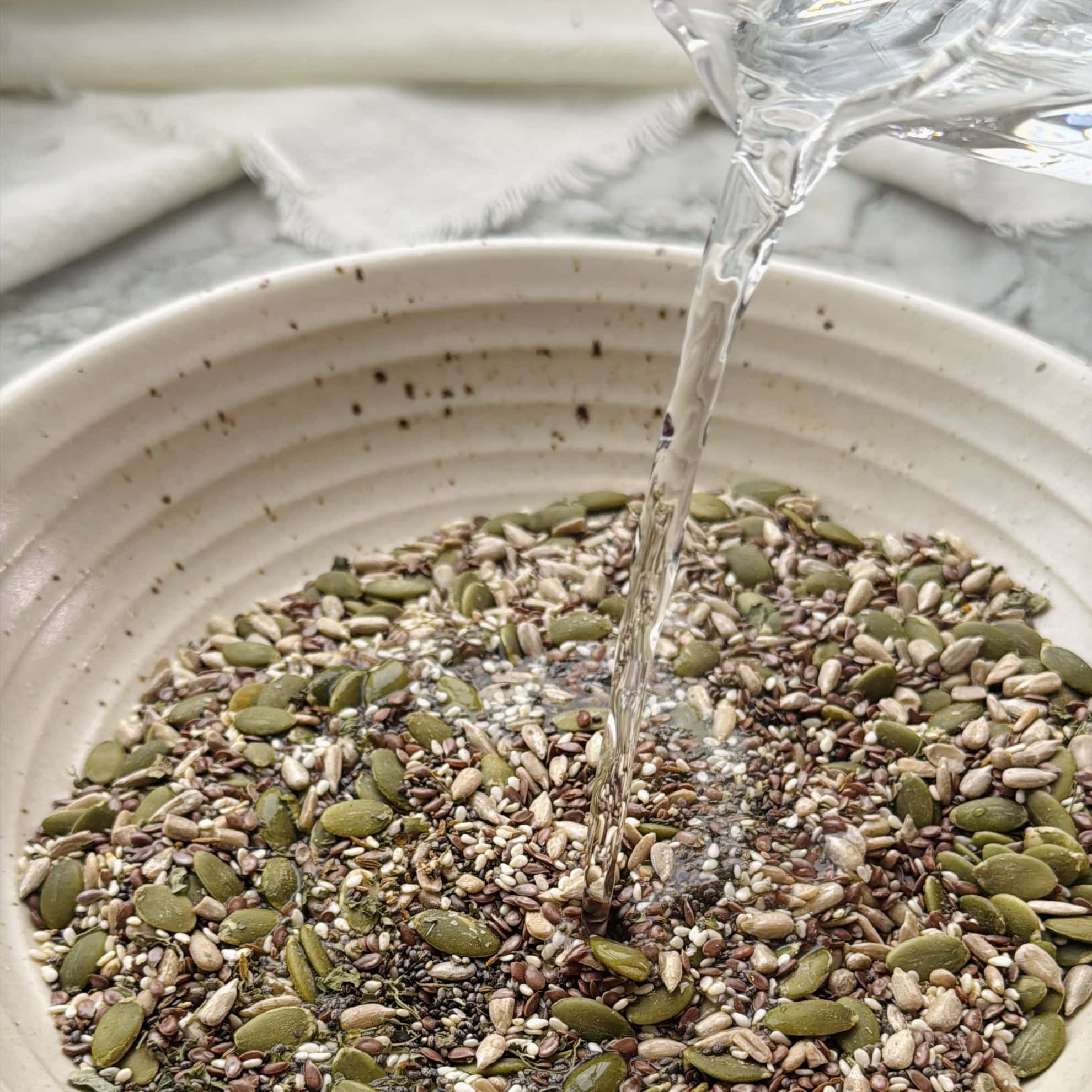 Water is being poured into a bowl filled with a mixture of seeds, including pumpkin, sunflower, sesame, and flax seeds, on a marble surface with a cloth in the background.