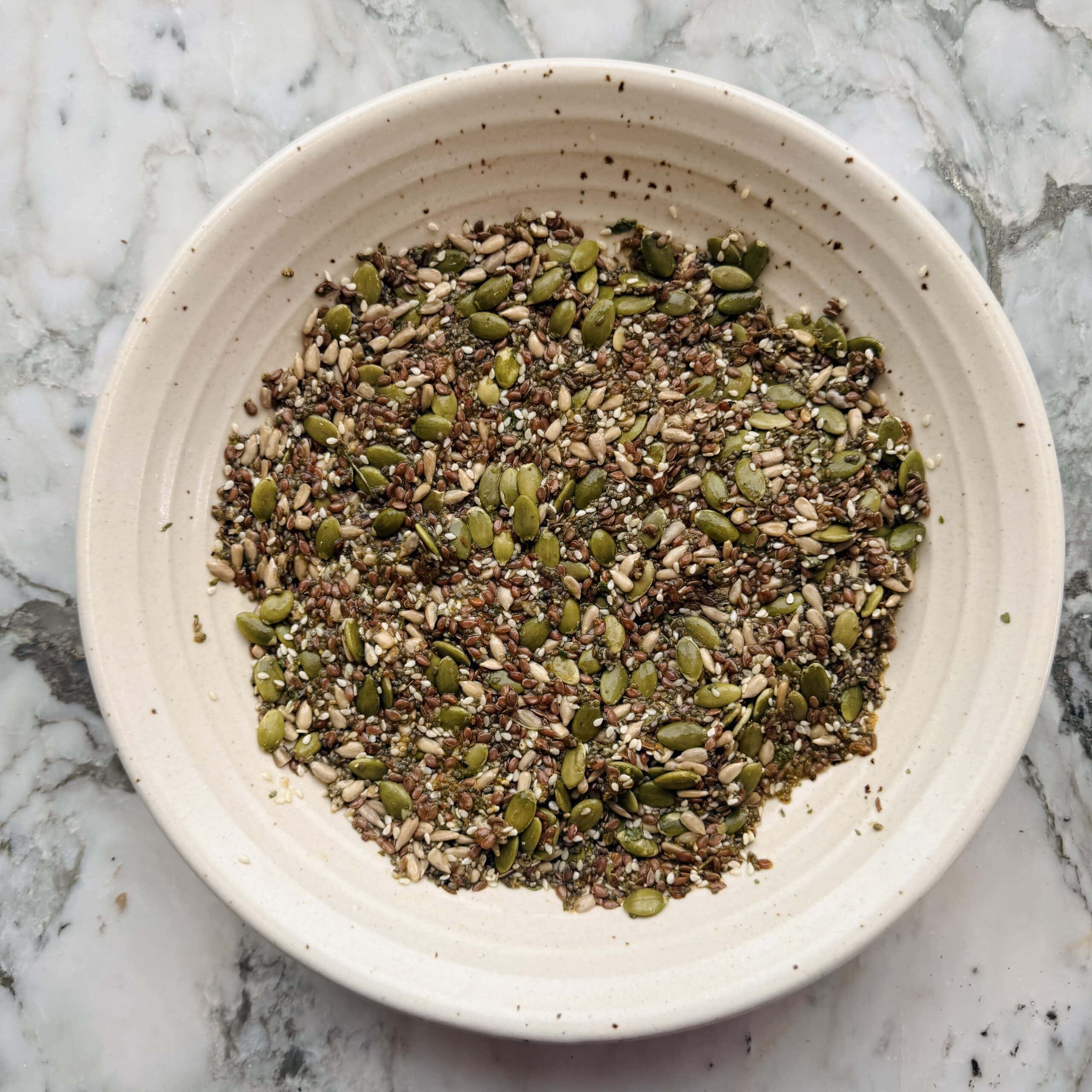 A beige bowl containing a mixture of seeds, including pumpkin, sunflower, flax, and sesame seeds, placed on a marble surface.