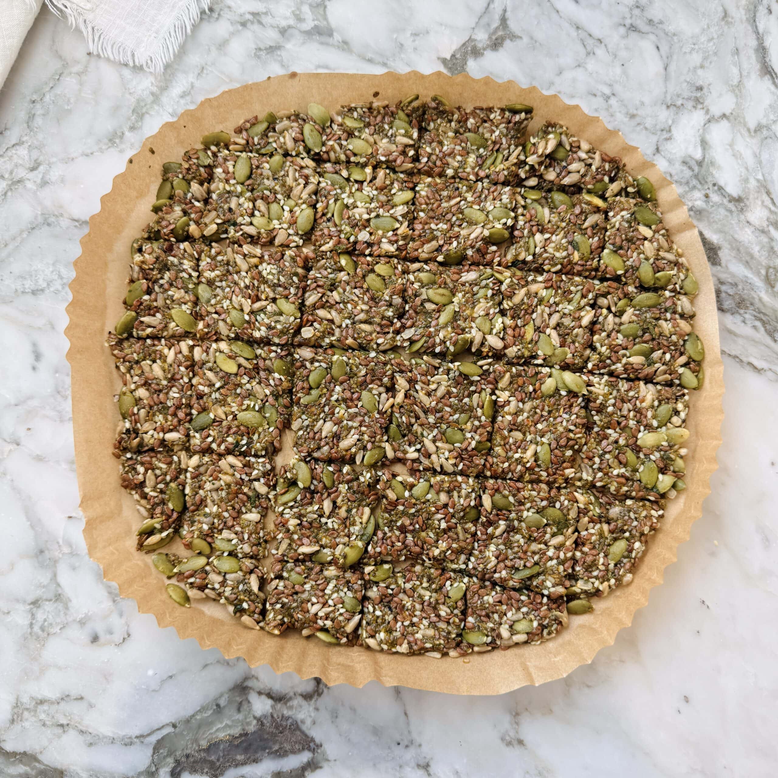Round tray of seed crackers, cut into squares, sits on parchment paper atop a marble surface. Crackers contain mixed seeds, including pumpkin, sesame, and sunflower.