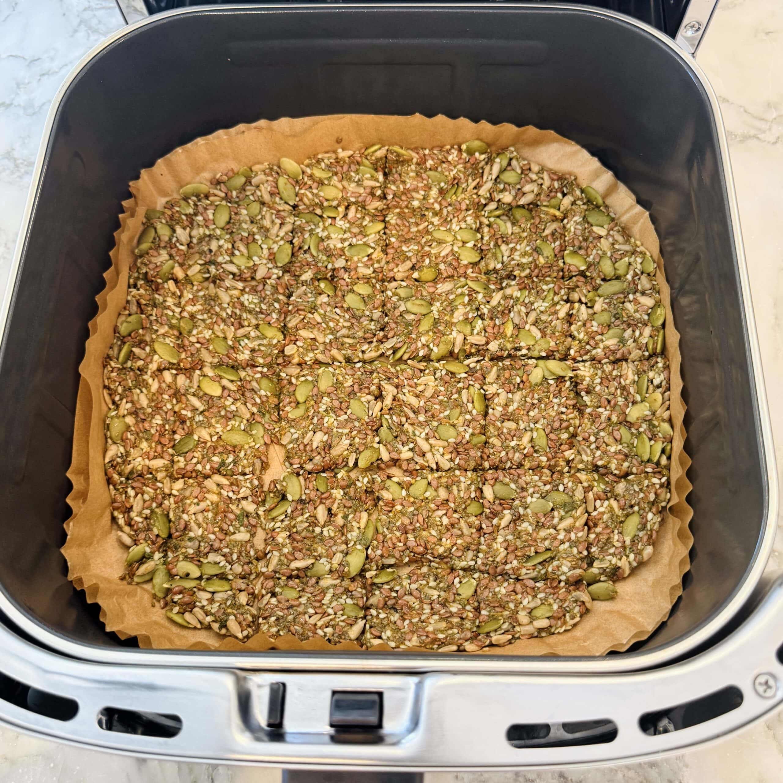 A sheet of seed crackers cut into squares sits on parchment paper inside an air fryer basket.