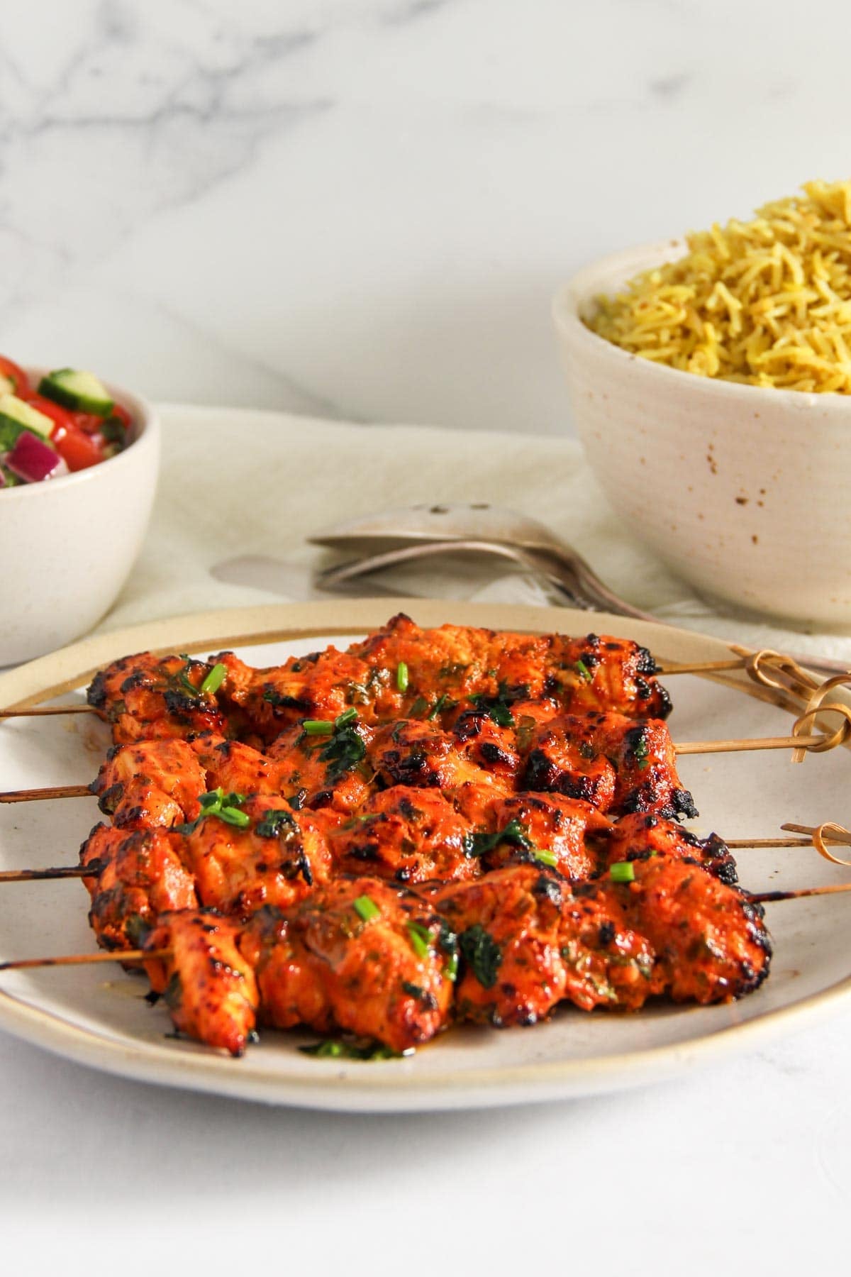 Grilled skewers of marinated chicken served on a plate, accompanied by a bowl of yellow rice and a bowl of salad in the background.