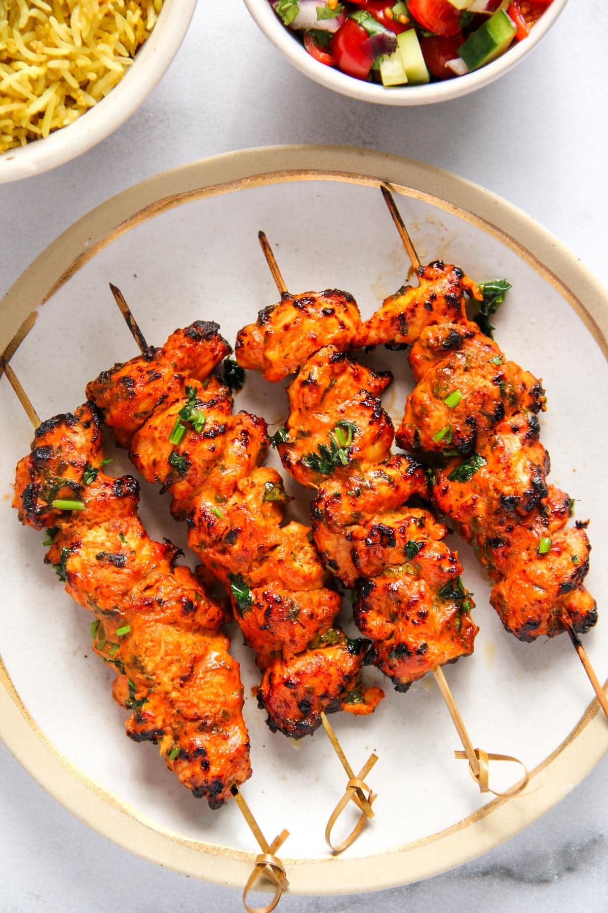 Four skewers of grilled, marinated chicken kebabs garnished with herbs are arranged on a round white plate, with bowls of rice and salad nearby.