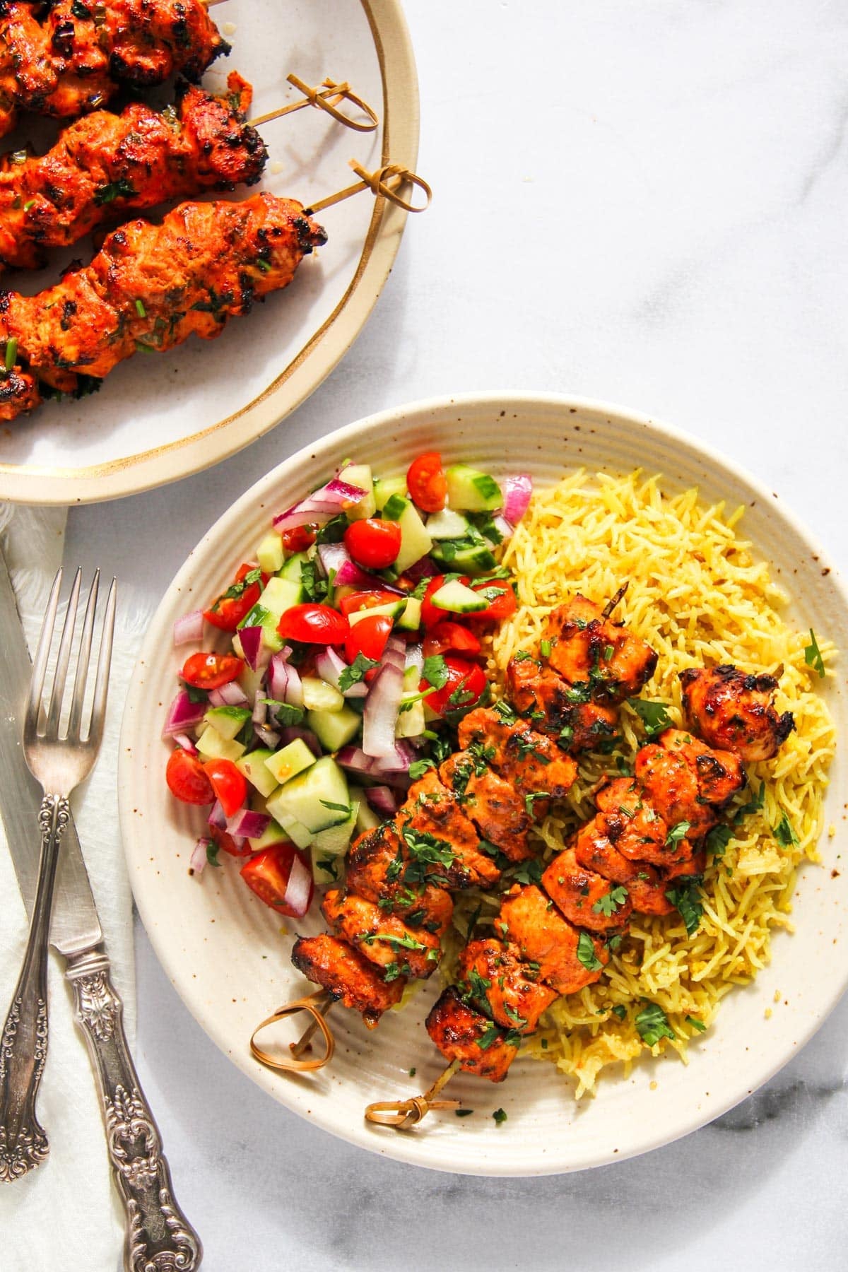 A plate with grilled chicken skewers, yellow rice, and a side of chopped cucumber, tomato, and red onion salad. Another plate of skewers is partially visible above.