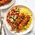 A plate with two grilled chicken skewers, yellow rice, and a side of chopped cucumber, tomato, and red onion salad. Fork and knife placed to the left.