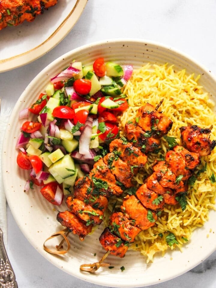 A plate with two grilled chicken skewers, yellow rice, and a side of chopped cucumber, tomato, and red onion salad. Fork and knife placed to the left.